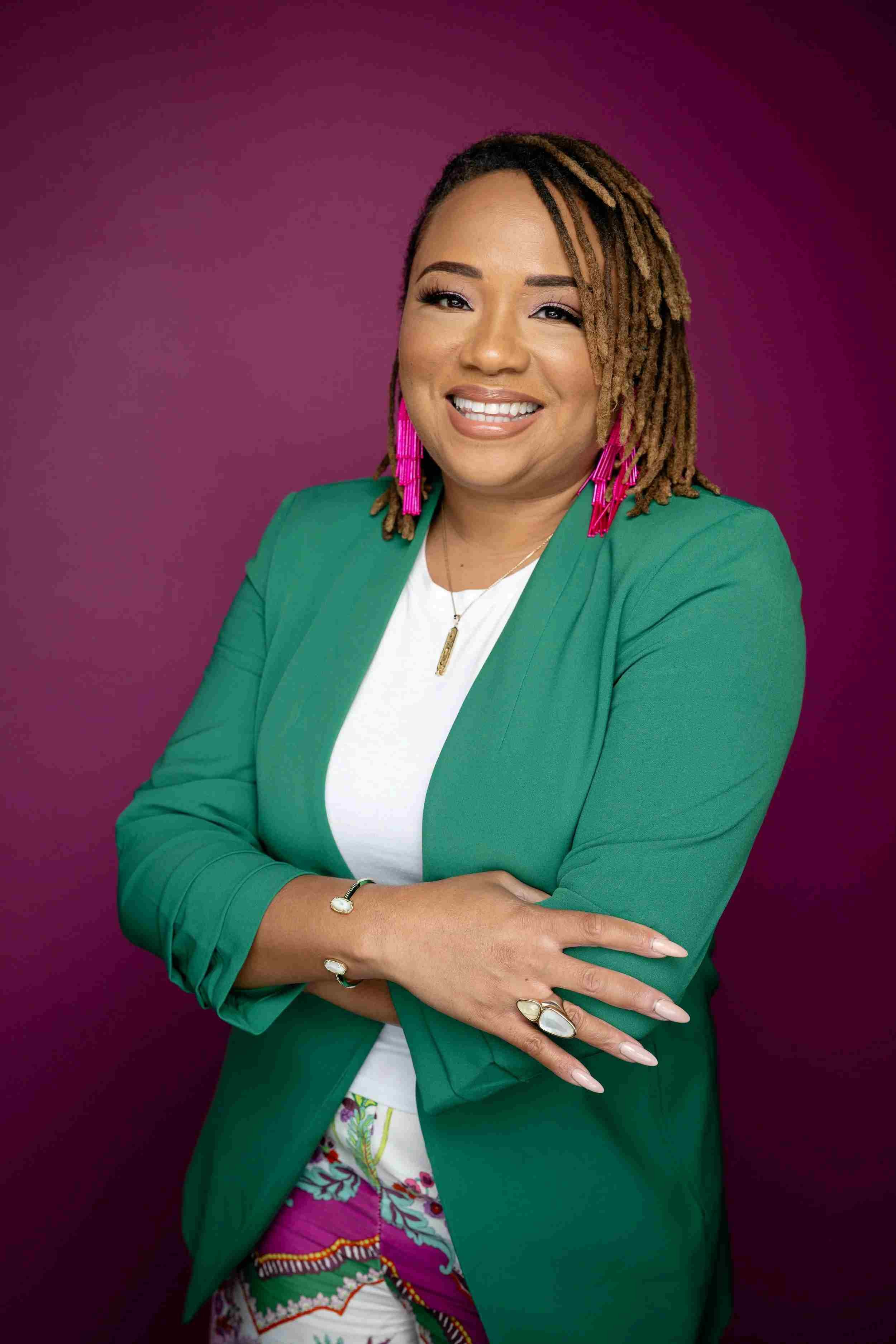 Portrait of a woman with dreadlocks, wearing a green blazer, white top, and colorful patterned pants, standing with arms crossed in front of a purple background.