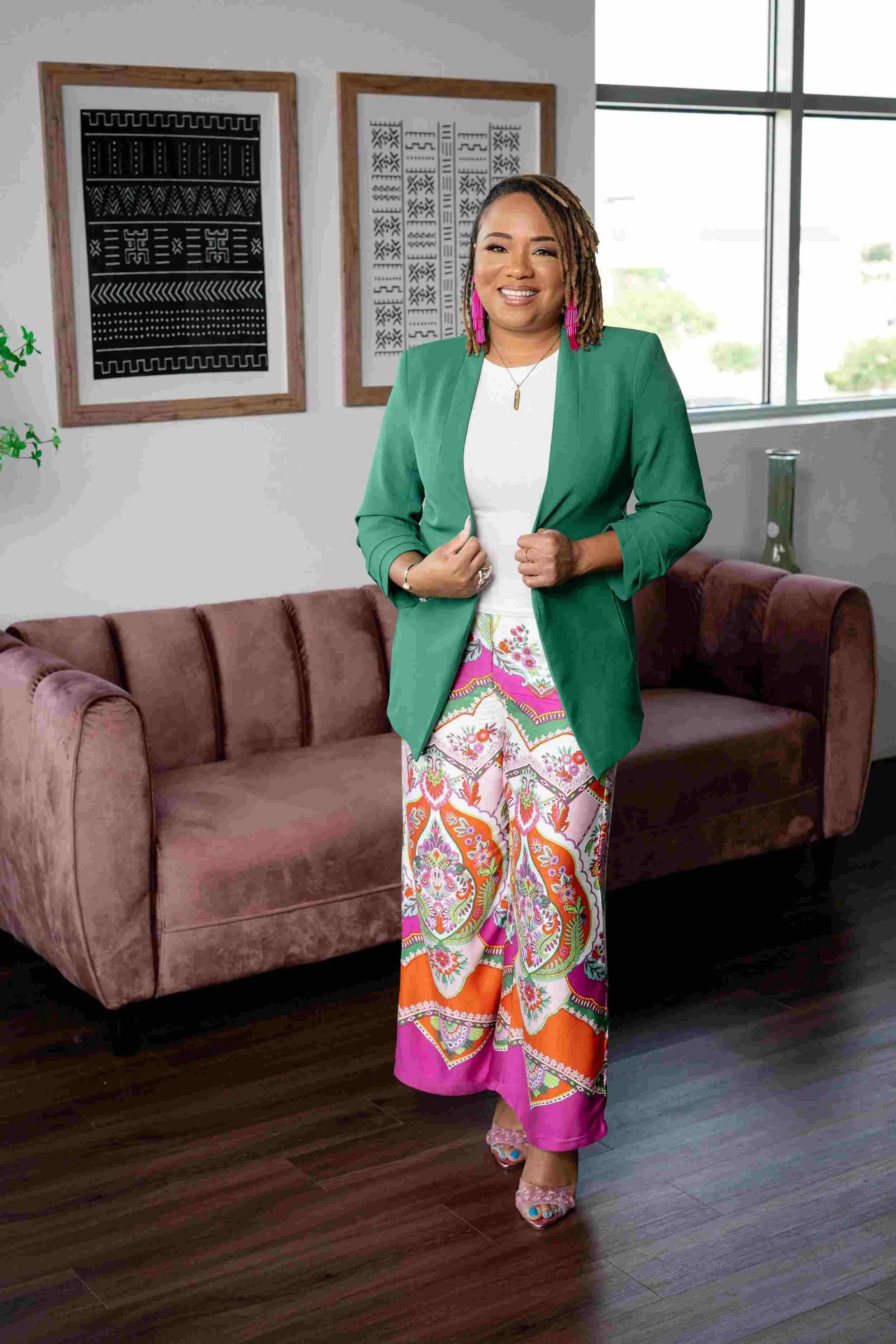 A woman with medium-length dreadlocks and pink earrings, wearing a green blazer, white top, colorful patterned pants, and open-toed sandals, standing in a modern living room with a pink velvet sofa, framed black and white artwork, and a window with a view of trees outside.