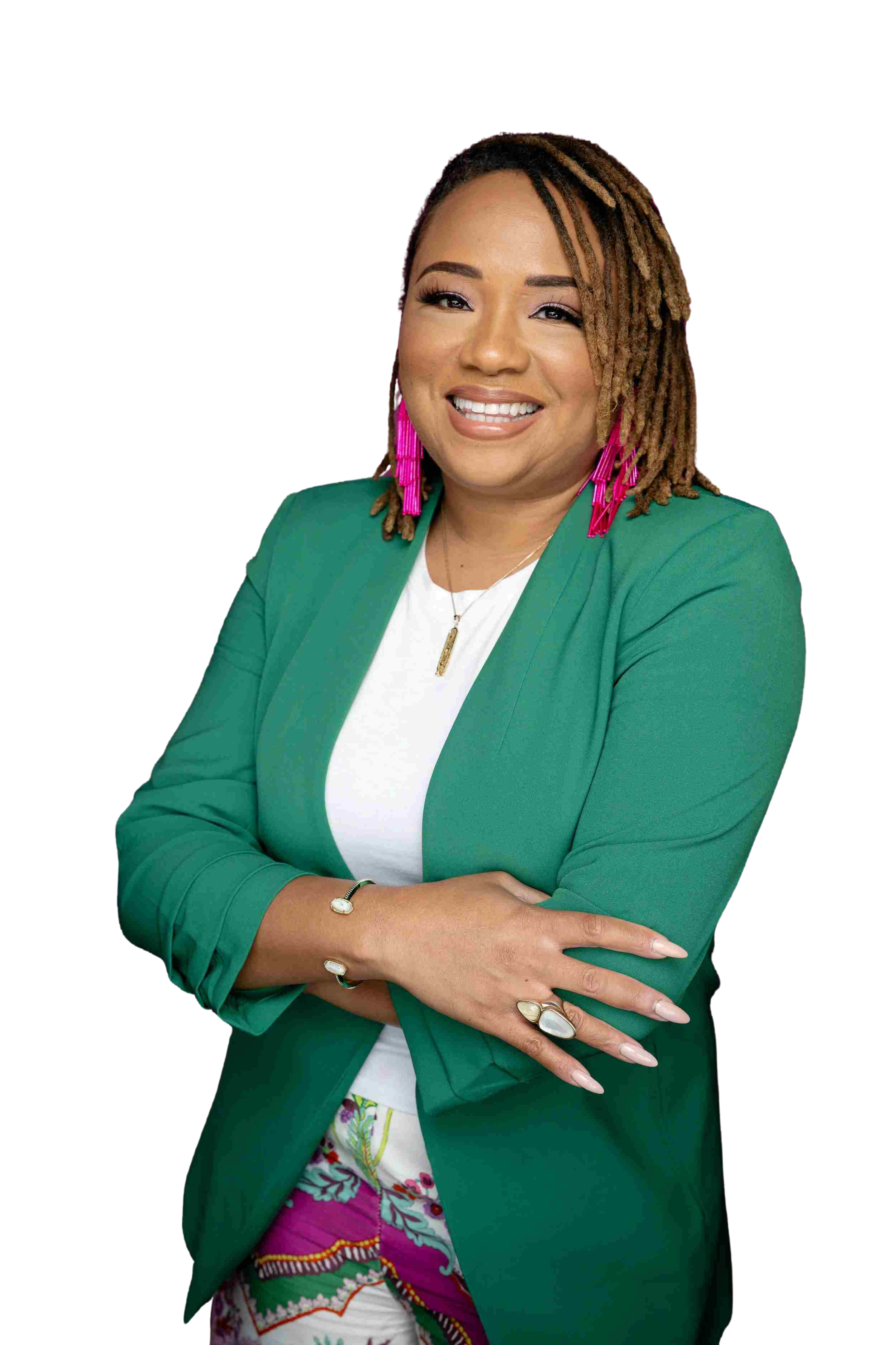 A woman with shoulder-length dreadlocks, wearing a green blazer, white top, and colorful patterned pants, smiling with crossed arms, accessorized with pink earrings, a gold necklace, and rings.