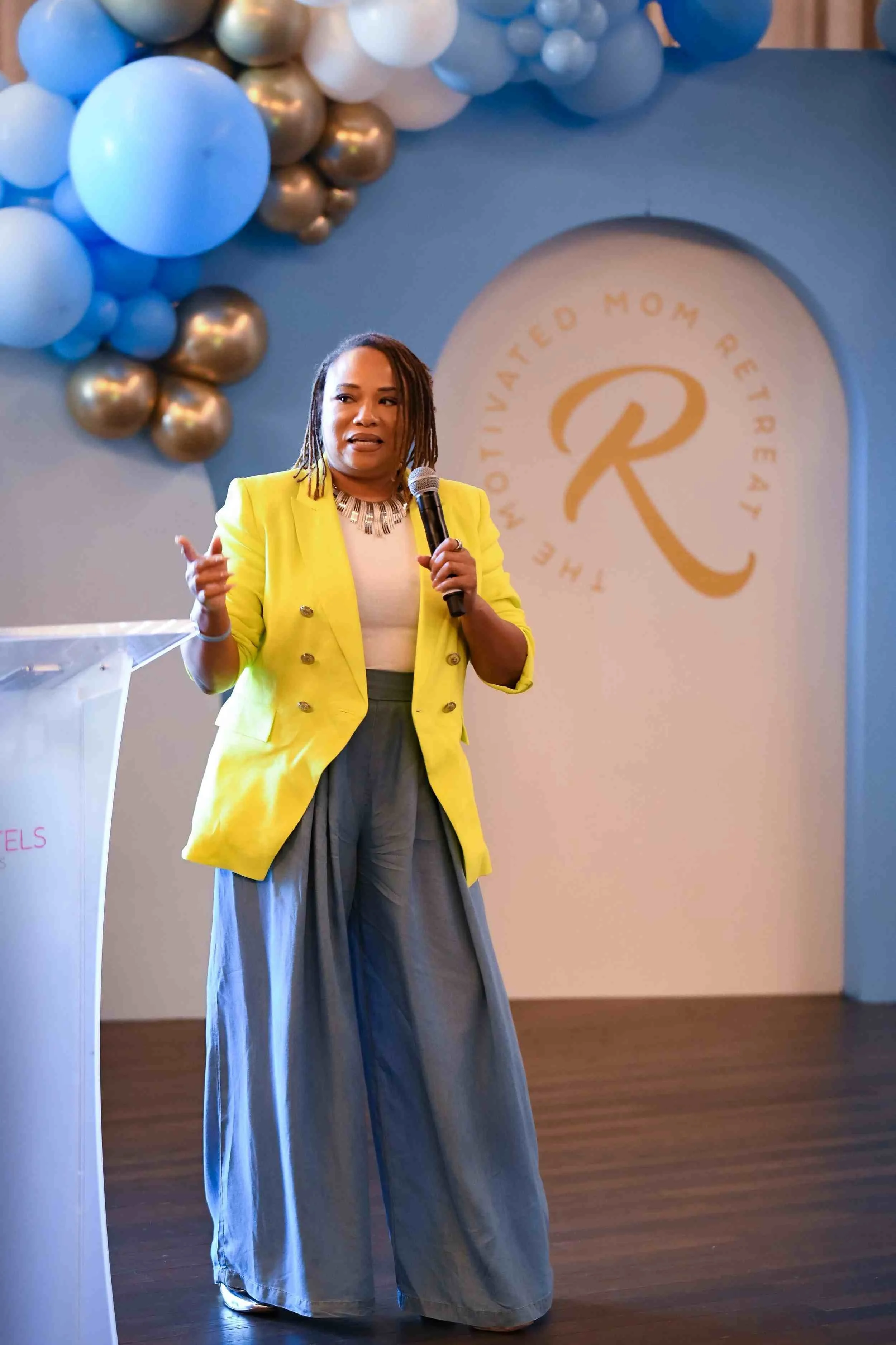A woman in a bright yellow blazer speaking into a microphone at an event with a backdrop that reads "The Motivated Mom Retreat" and features a logo with the letter "R". She is wearing a white top and grey wide-leg pants, standing on a wooden floor, with balloons in white, blue, and gold above her.