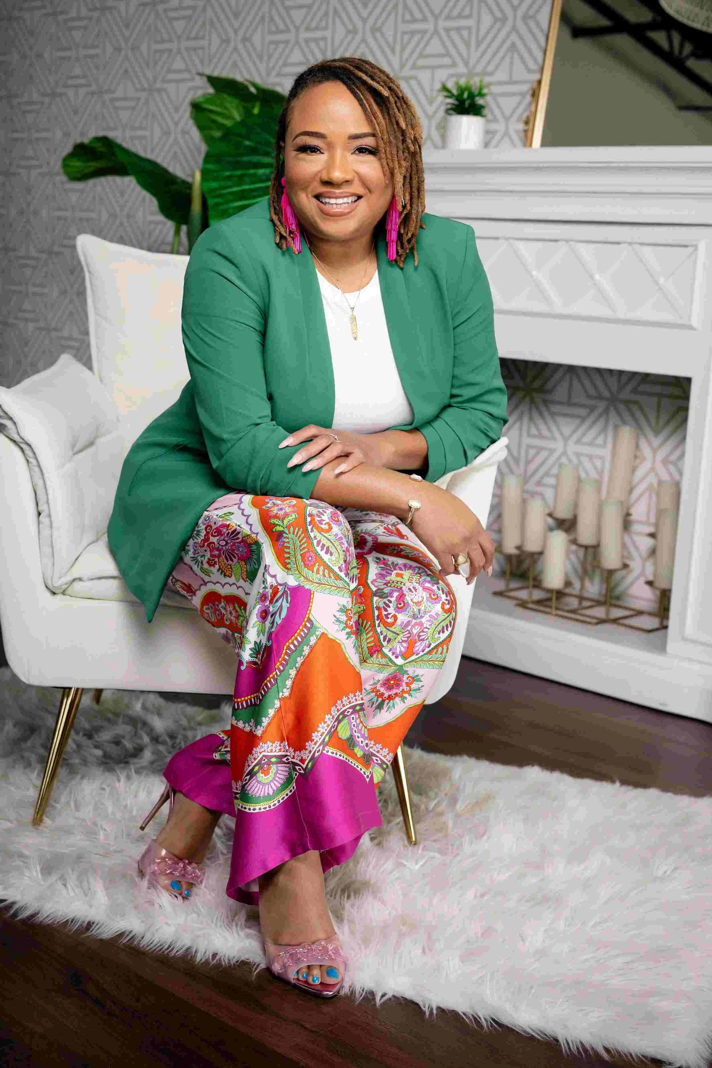 A woman sitting on a white chair, smiling, wearing a green blazer, white top, colorful paisley-patterned pants, and pink heels, with decor including plants, candles, and geometric wallpaper in the background.