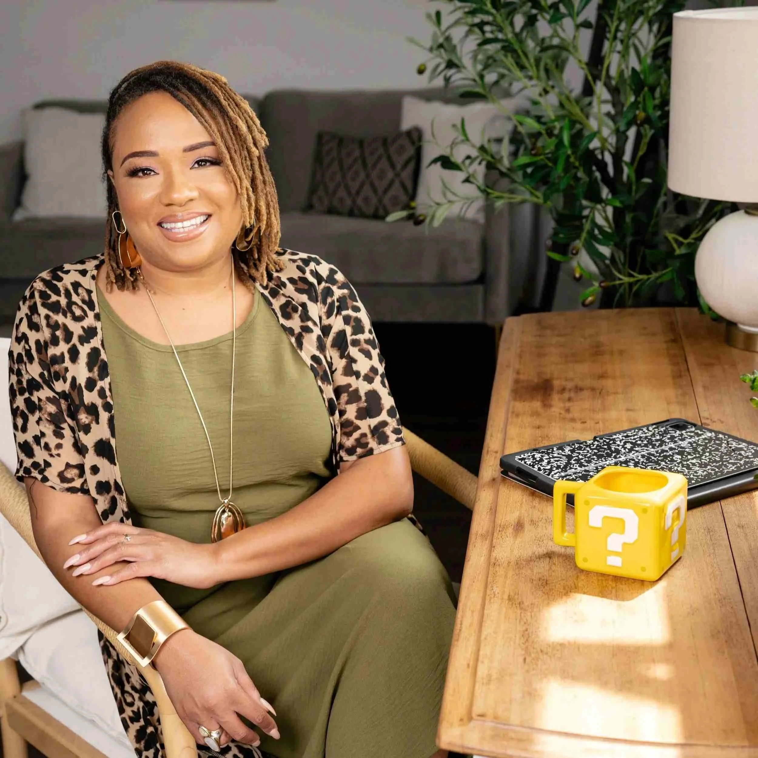 A woman with short, blonde dreadlocks sitting at a wooden table in a living room, smiling at the camera. On the table, there is a black and white patterned device, a yellow question mark block, and a smartphone. Behind her, there is a gray sofa with patterned pillows, a large green plant, and a white table lamp.