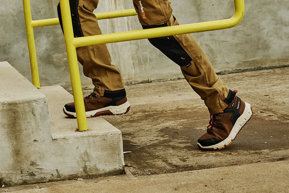 Close-up of person wearing brown pants and Timberland hiking boots, leaning on a yellow handrail near concrete stairs on a sidewalk.