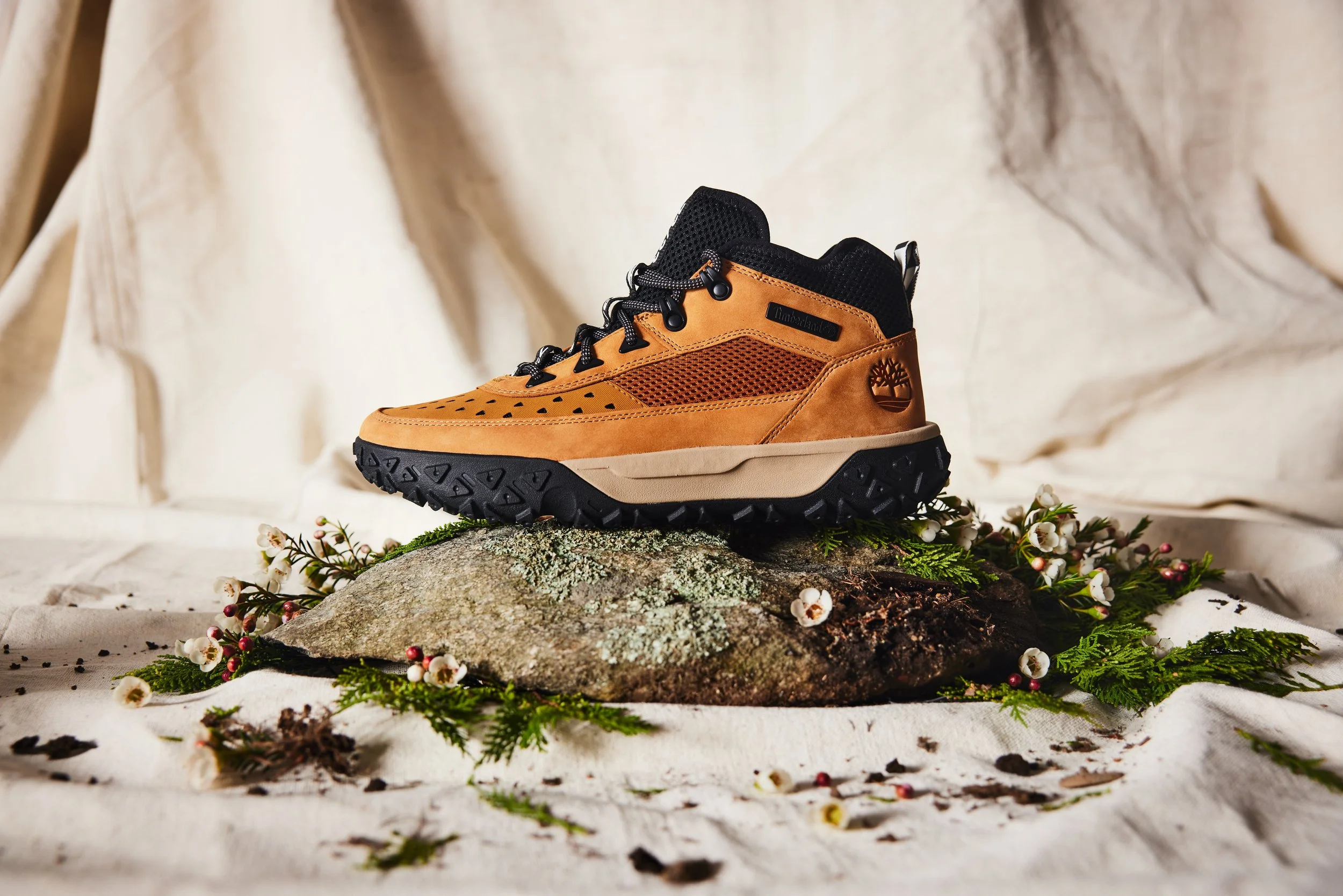 A tan hiking shoe with black accents and rugged sole, placed on a mossy rock surrounded by evergreen branches, white flowers, and a white cloth backdrop.