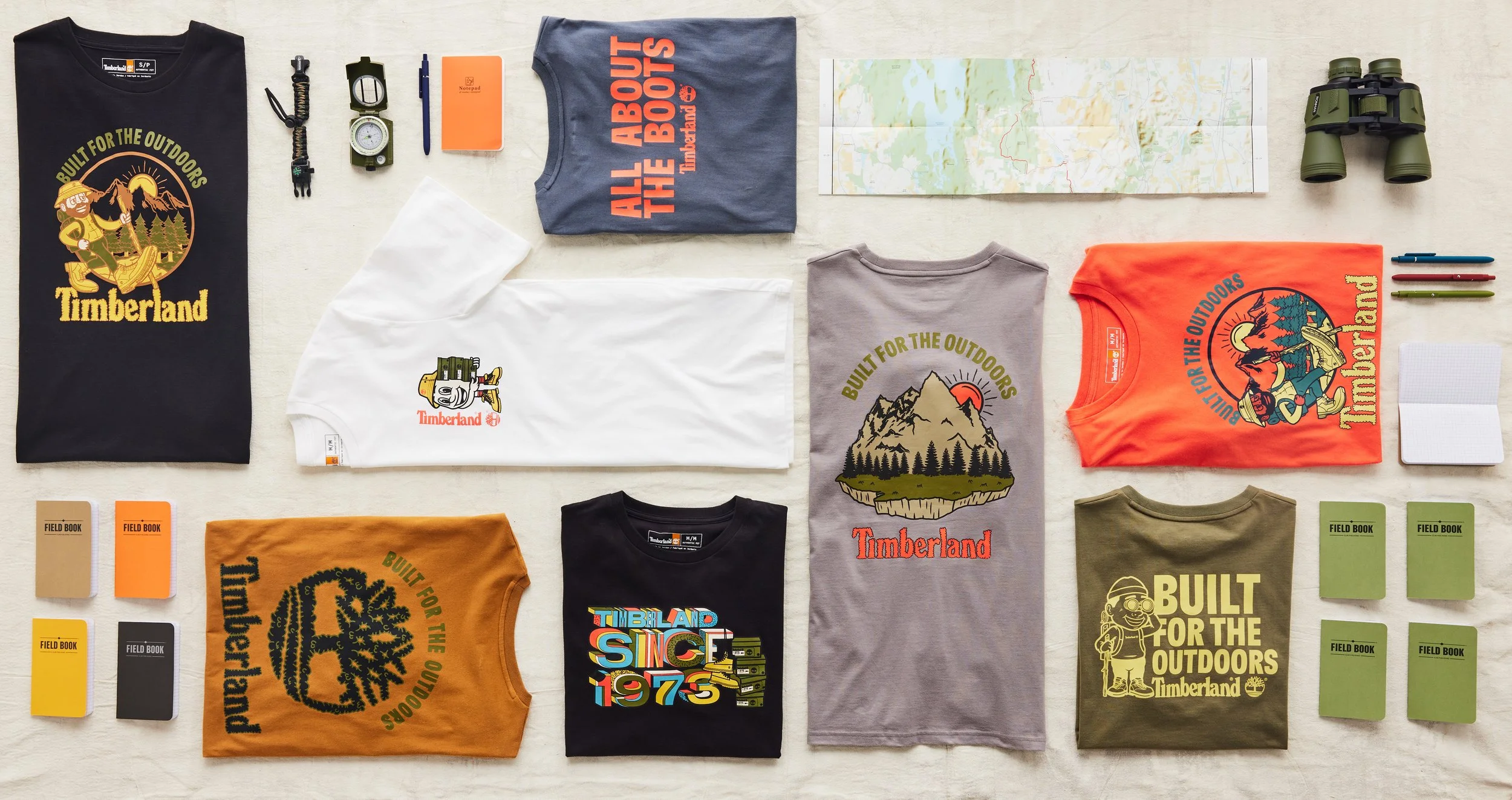 Flat lay of Timberland-themed apparel and accessories including T-shirts with outdoor and mountain designs, notebooks, pens, a map, binoculars, a watch, a flashlight, a whistle, and a small speaker, displayed on a light-colored surface.