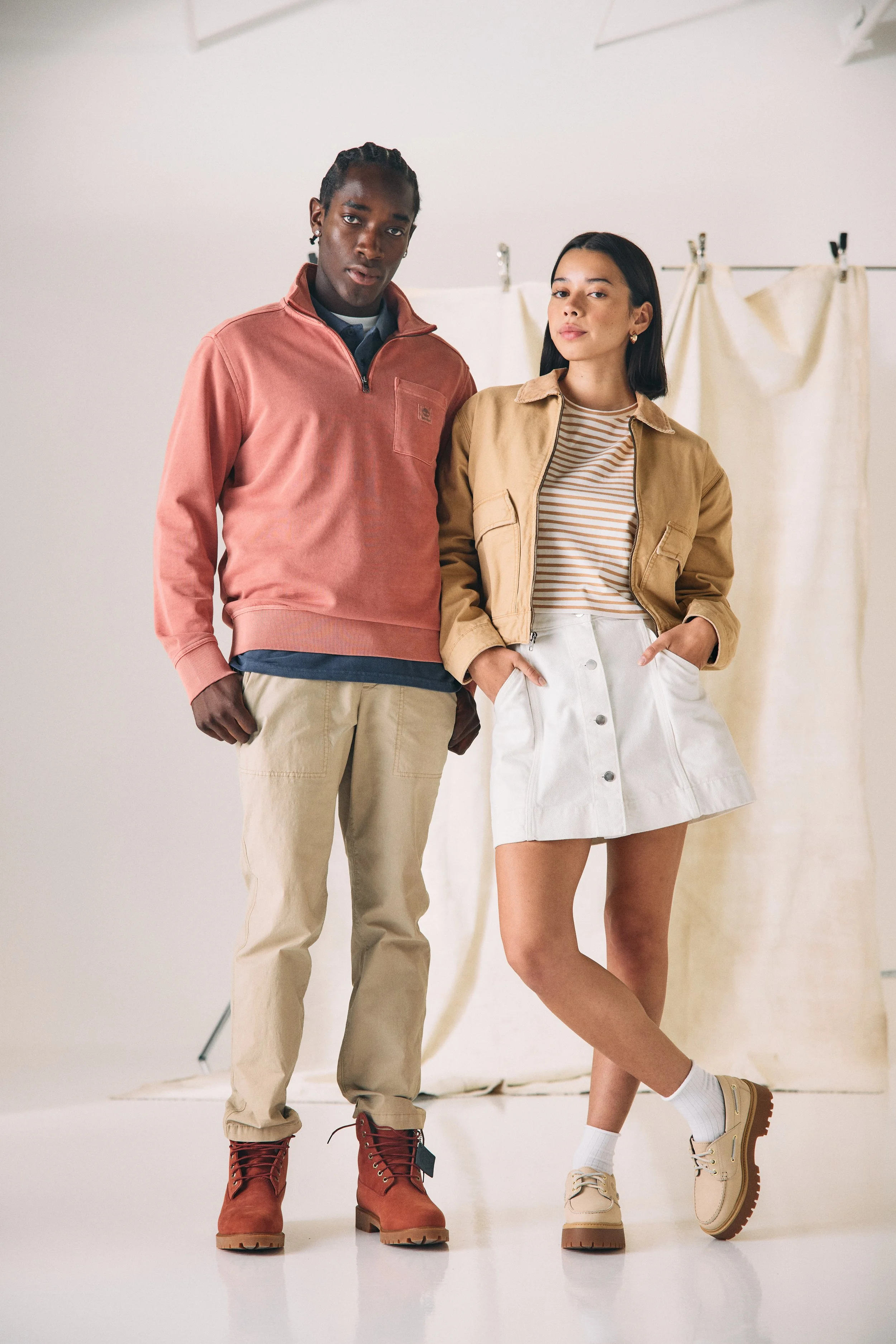 A man and woman posing in casual outfits in a studio with a cream backdrop.