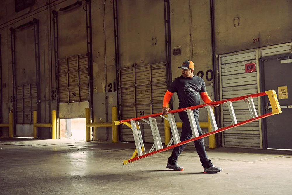 A man walking inside a warehouse carrying a yellow and red ladder.