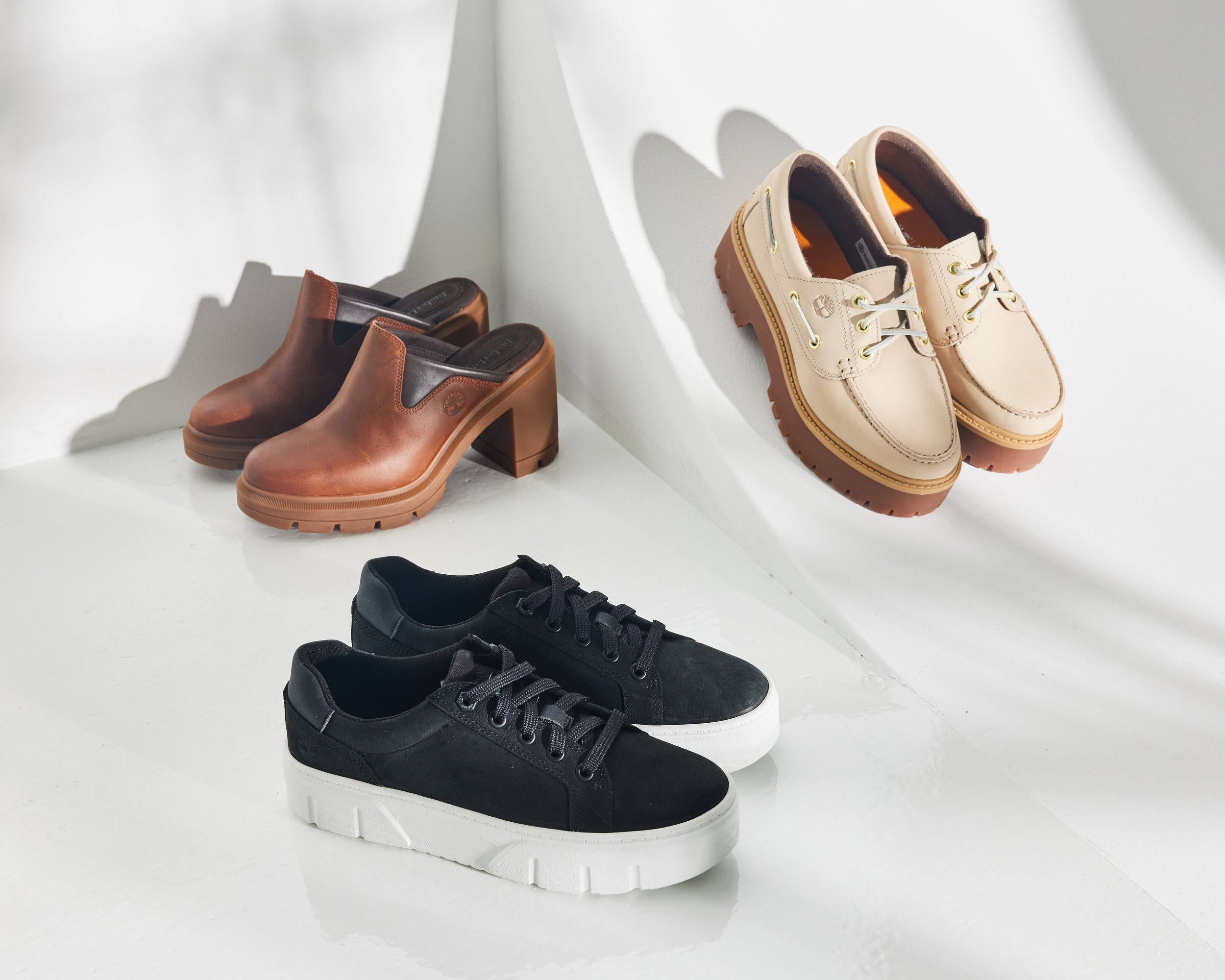 Three pairs of women's Timberland shoes on a white surface: brown heeled slip-on boots, beige lace-up shoes, and black sneakers with white soles.