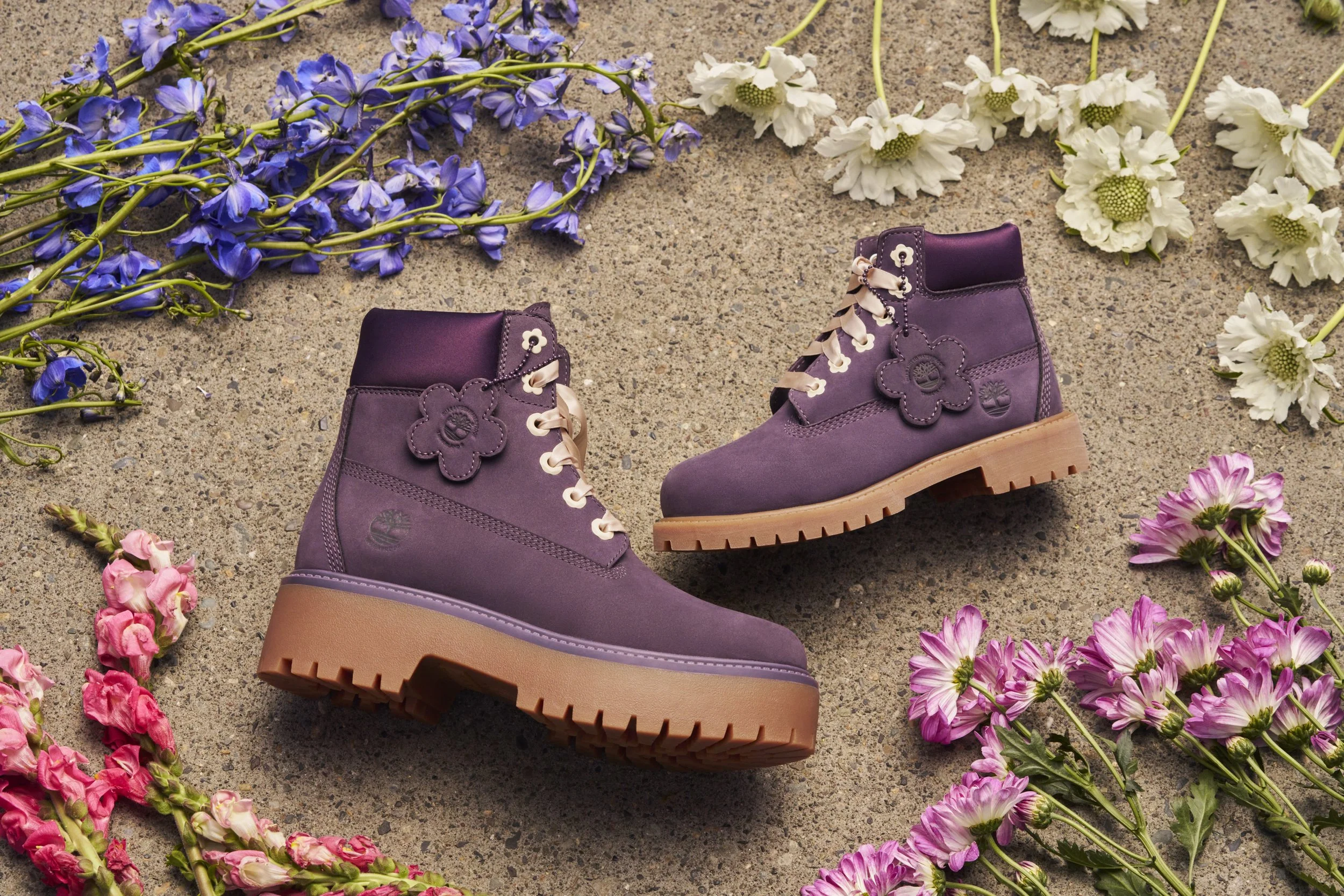 A pair of purple Timberland boots surrounded by pink, purple, and white flowers on a concrete surface.