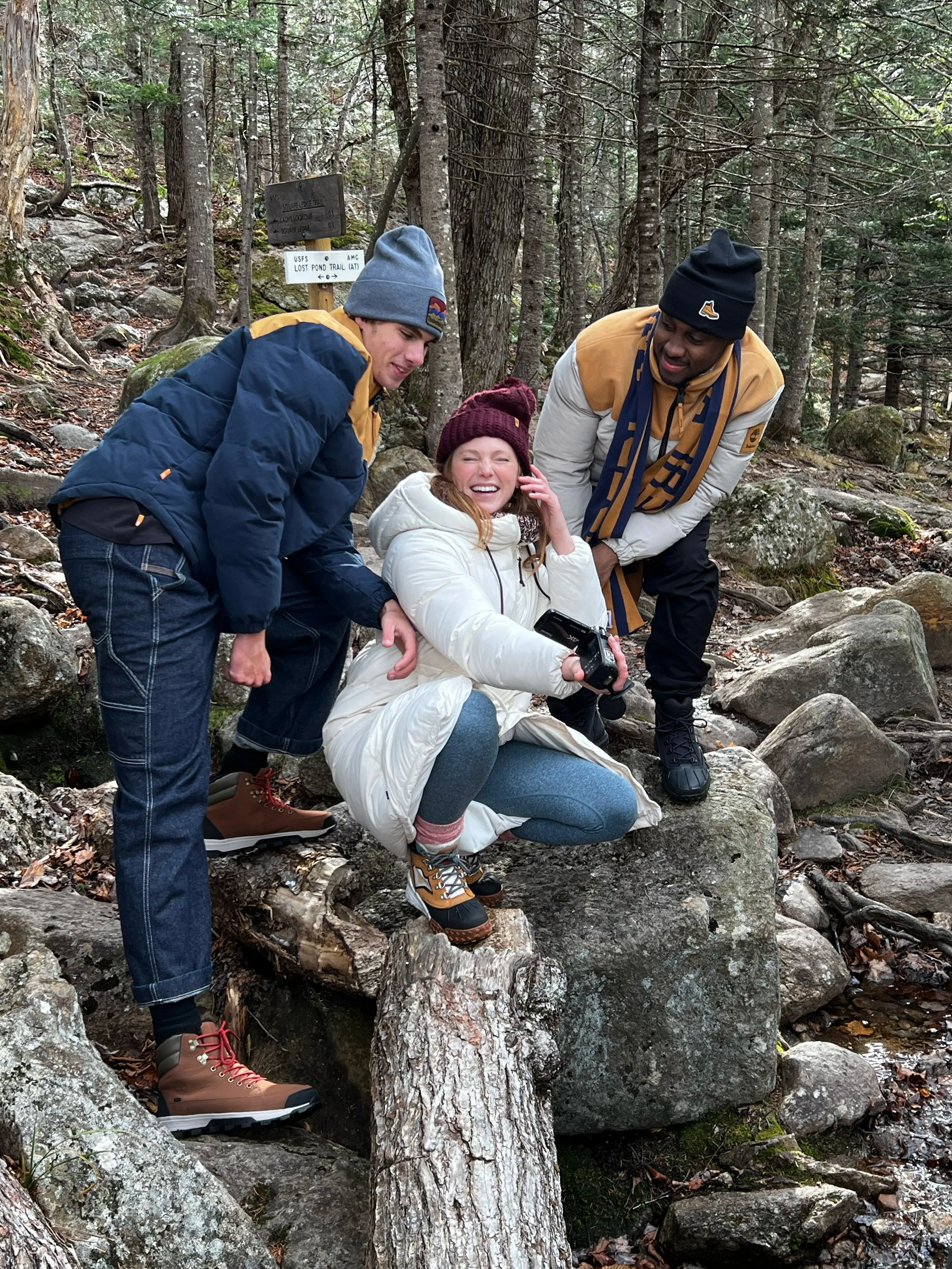 Three people in a forest, two men and one woman, are posing for a photo. The woman is sitting on a rock, smiling, and holding a camera. The men are standing on either side of her, leaning in, and smiling. They are dressed in outdoor clothing, includi