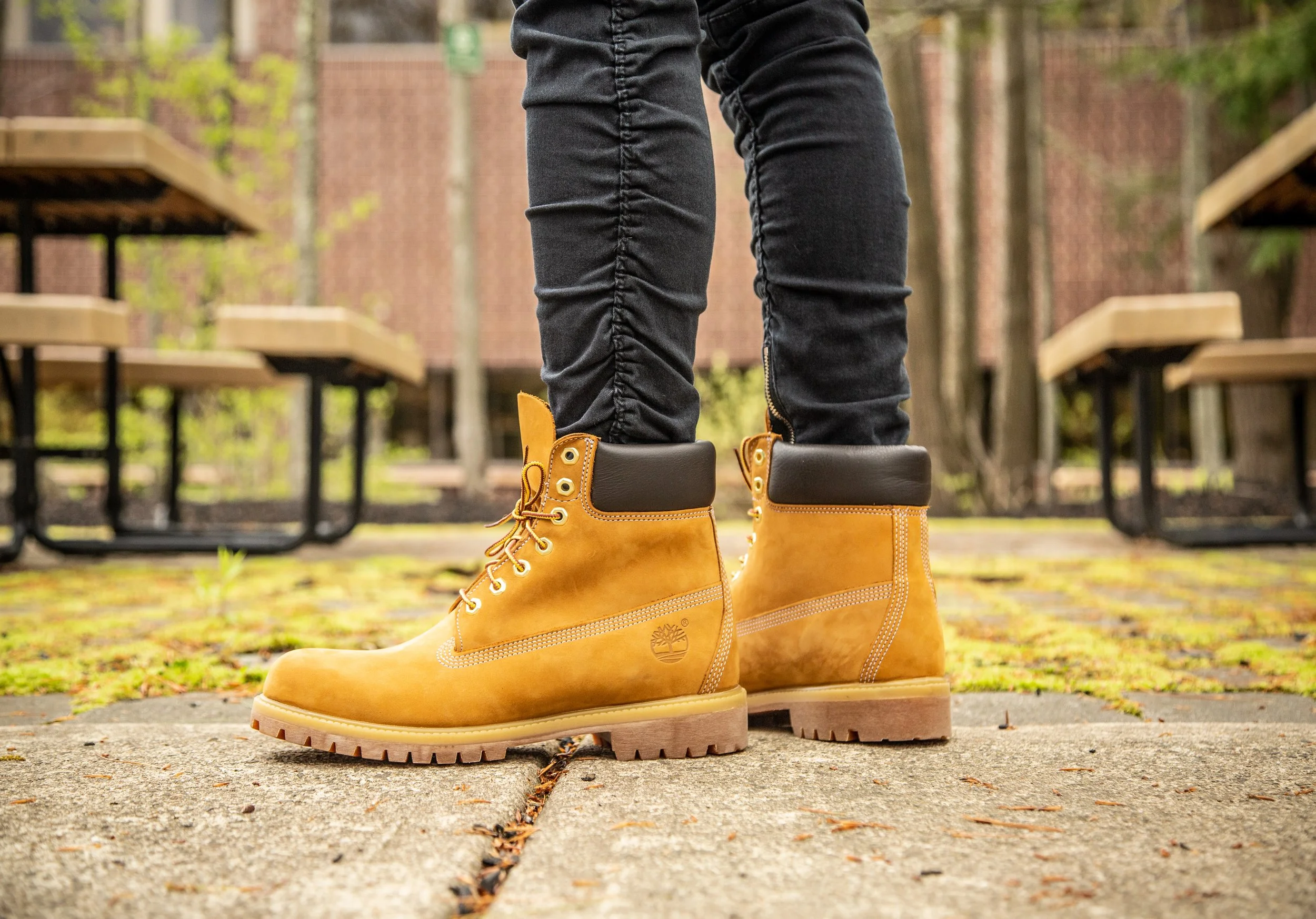 Close-up of person wearing yellow Timberland boots and dark jeans, standing on a concrete sidewalk in a park with benches and trees in the background.