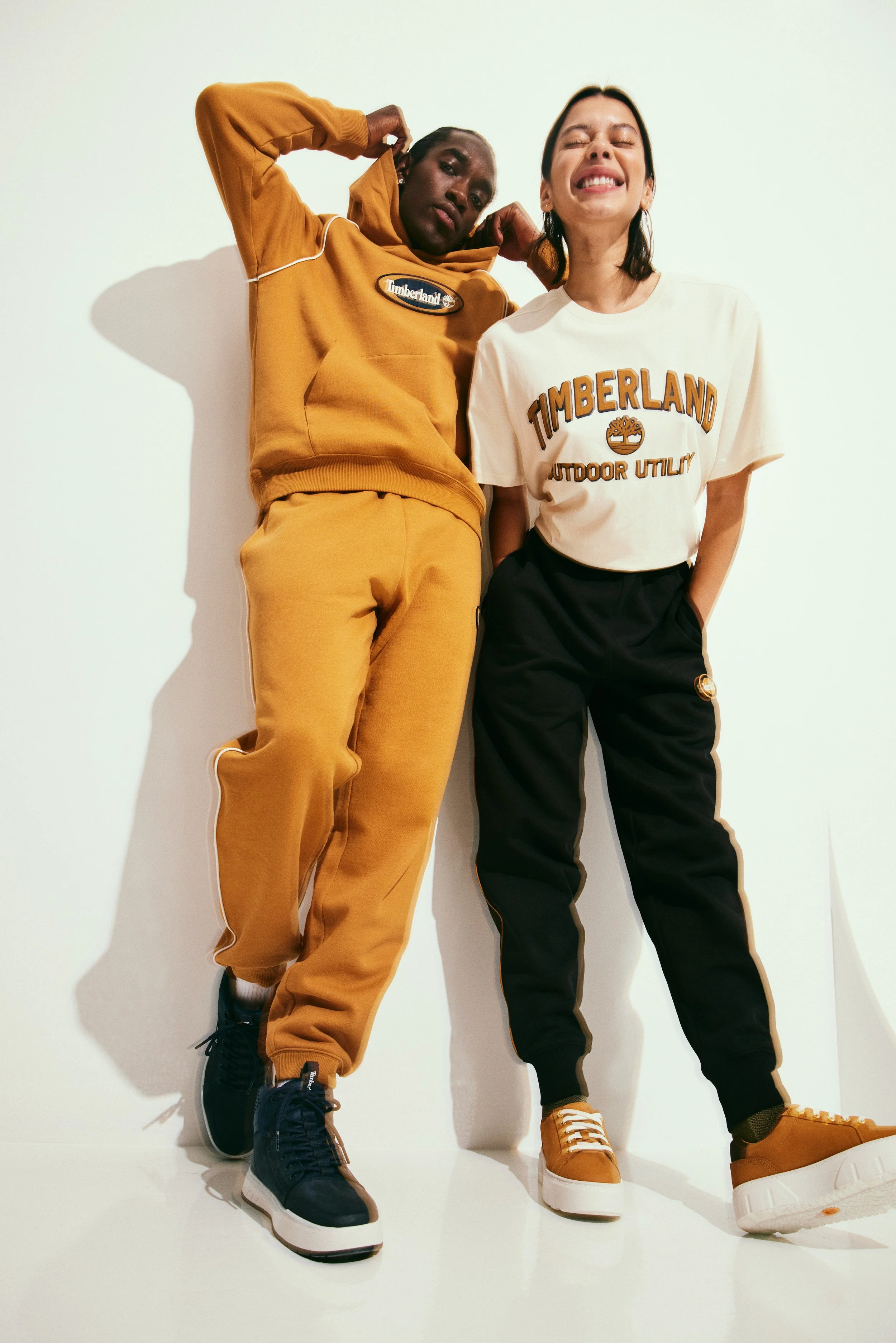 A man and a woman standing against a white wall, wearing Timberland casual clothing, with one in tan and the other in white and black, smiling and posing casually.