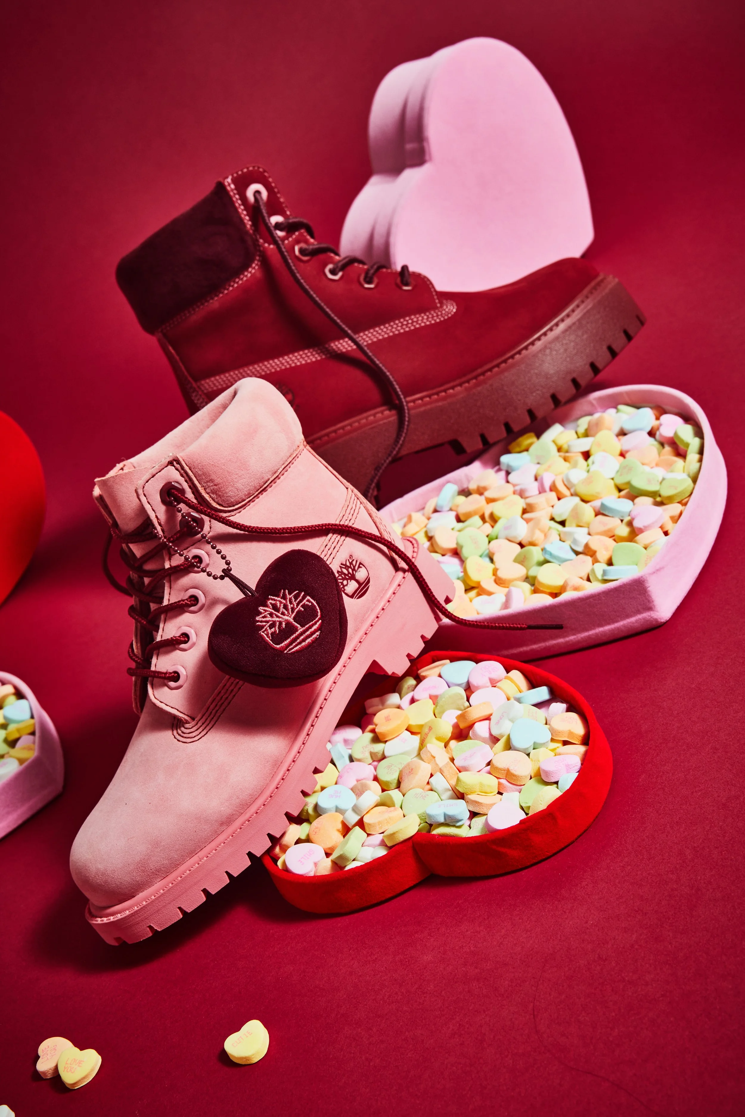 Pink and maroon Timberland boots surrounded by heart-shaped containers filled with pastel-colored candies, set on a red background for Valentine's Day.