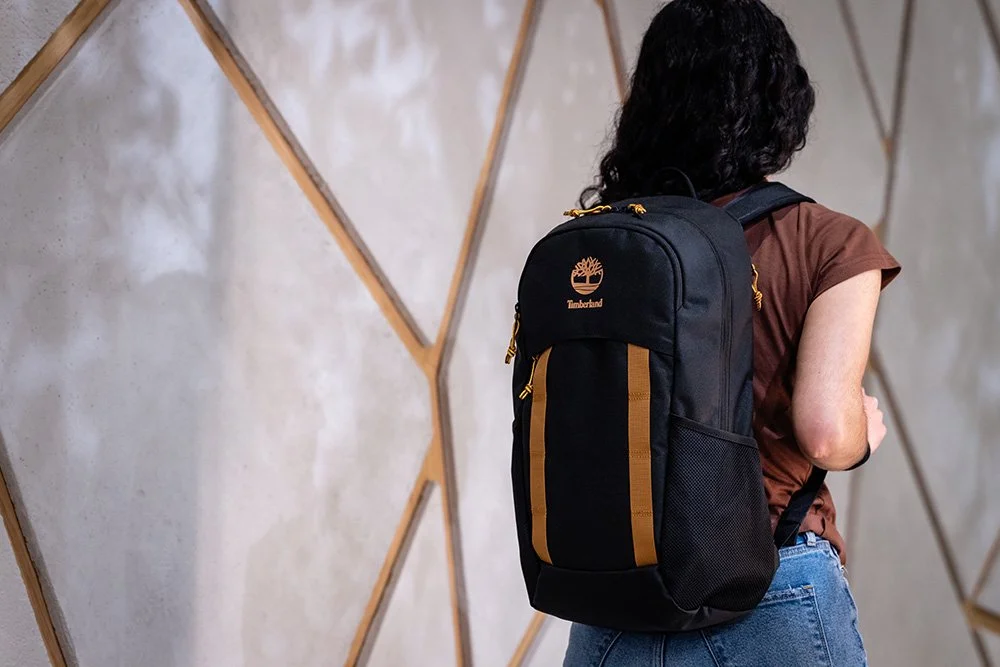A woman with curly black hair wearing a brown t-shirt and blue jeans carrying a black Timberland backpack with brown accents indoors against a patterned wall.