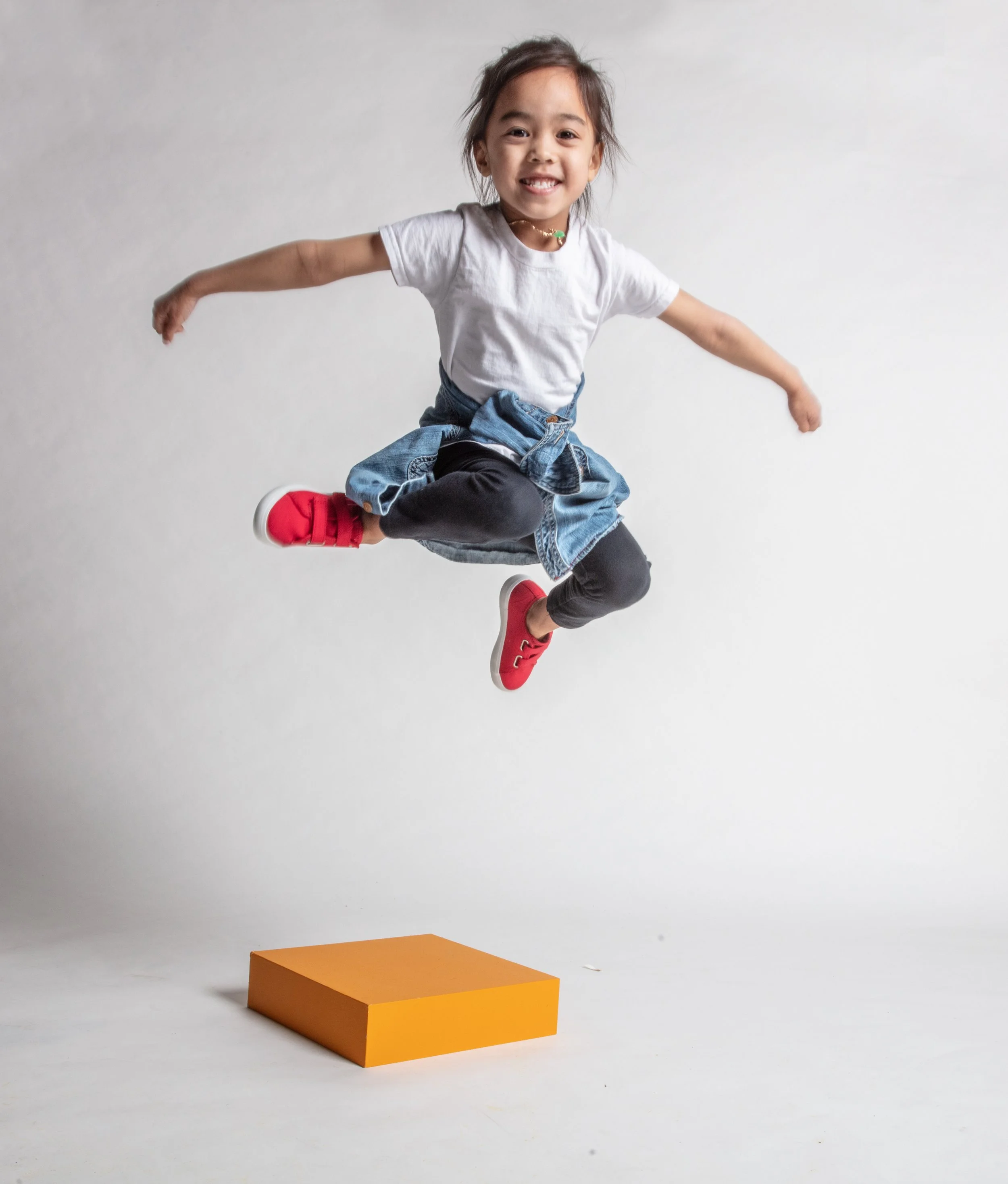 A young girl wearing a white t-shirt, black leggings, a denim jacket tied around her waist, and red sneakers jumping in the air over an orange box on a plain white background.