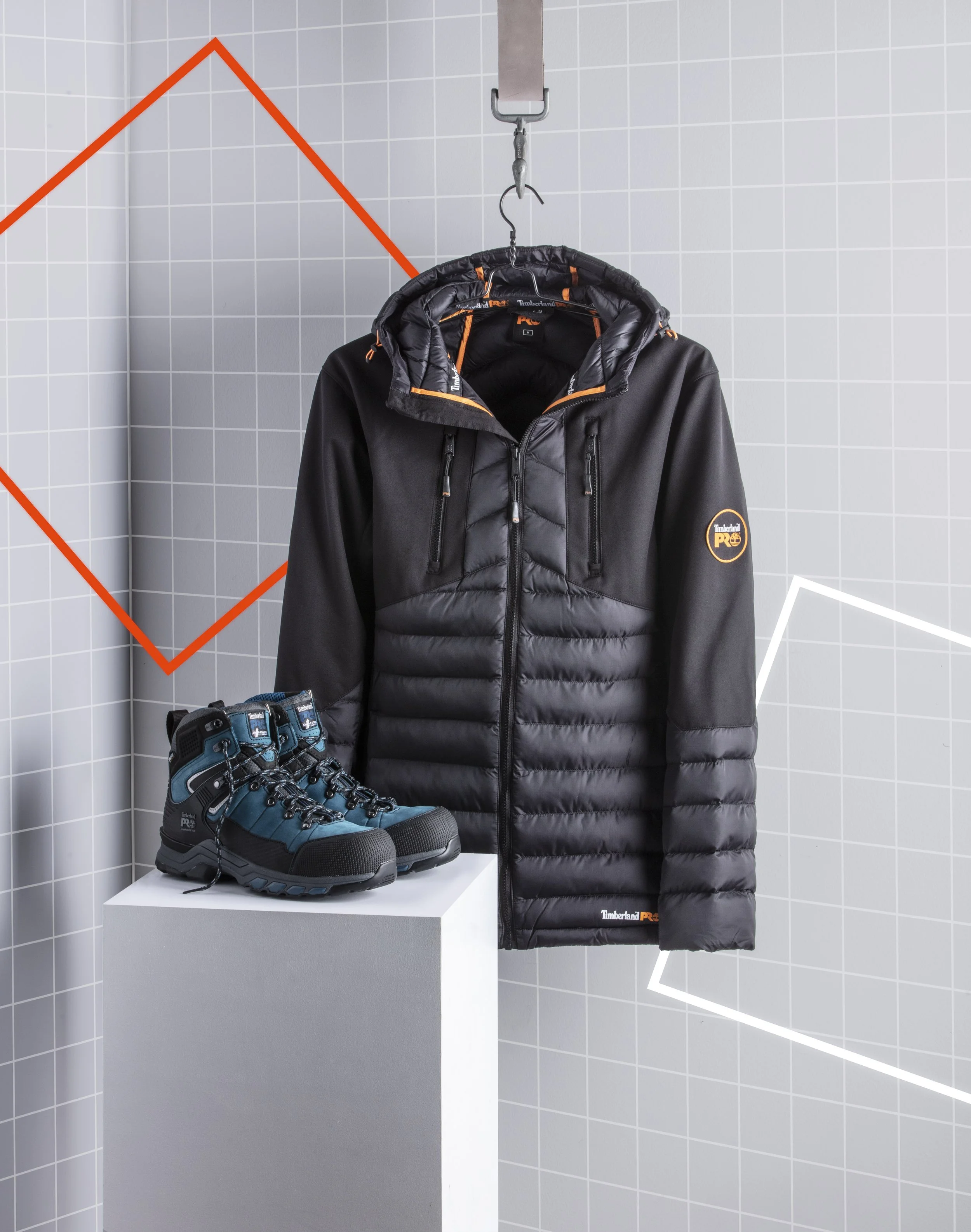 Black Timberland PRO winter jacket hanging on a hook and a pair of blue Timberland PRO outdoor boots displayed on a white pedestal against a grey tiled wall with geometric wall art.
