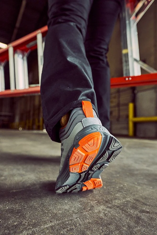 Close-up of a person walking in an industrial or warehouse setting, wearing gray and orange athletic shoes and black pants.