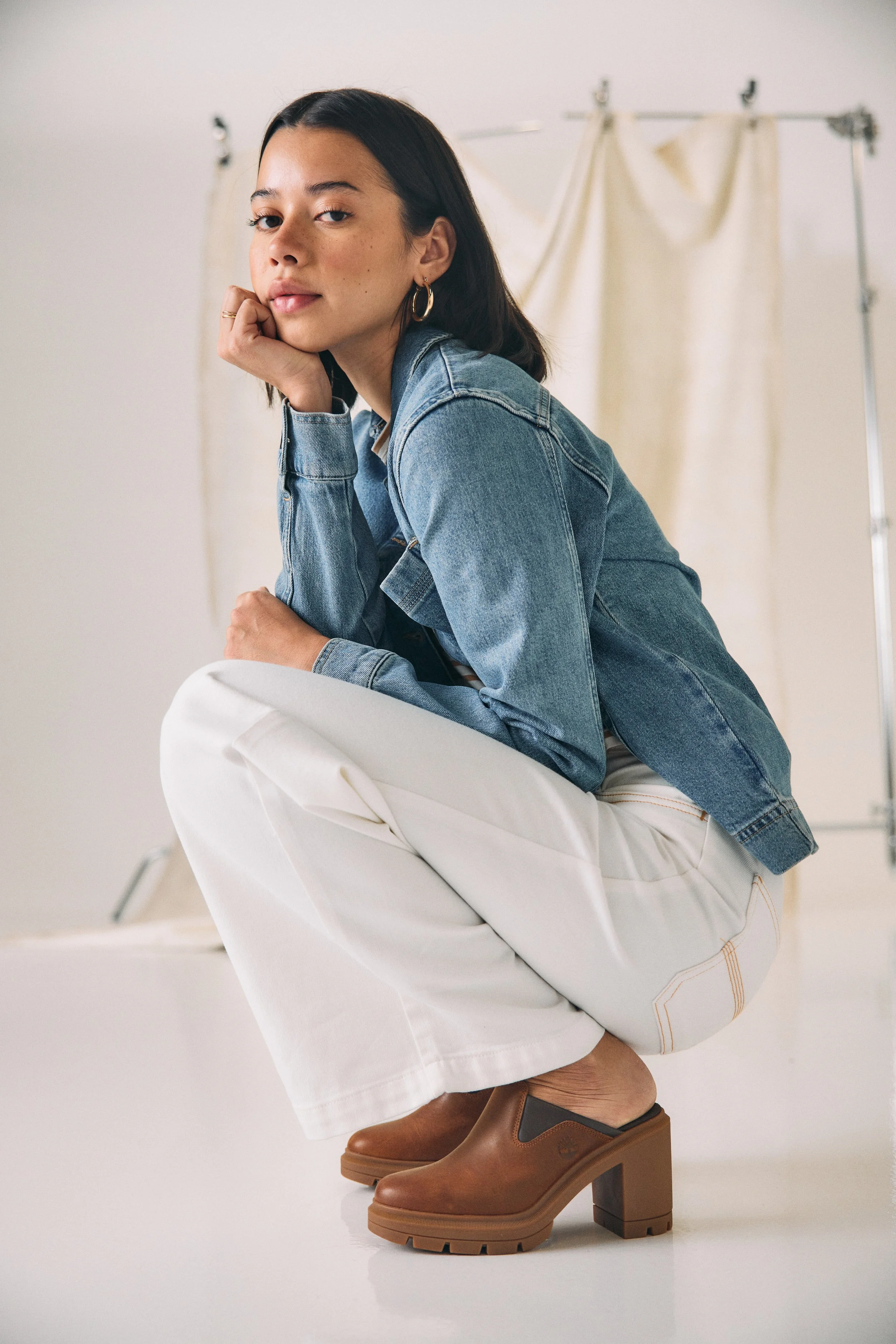 A woman crouched down, resting her chin on her hand, wearing a denim jacket, white pants, and brown chunky-heeled Timberland shoes, with a neutral background.
