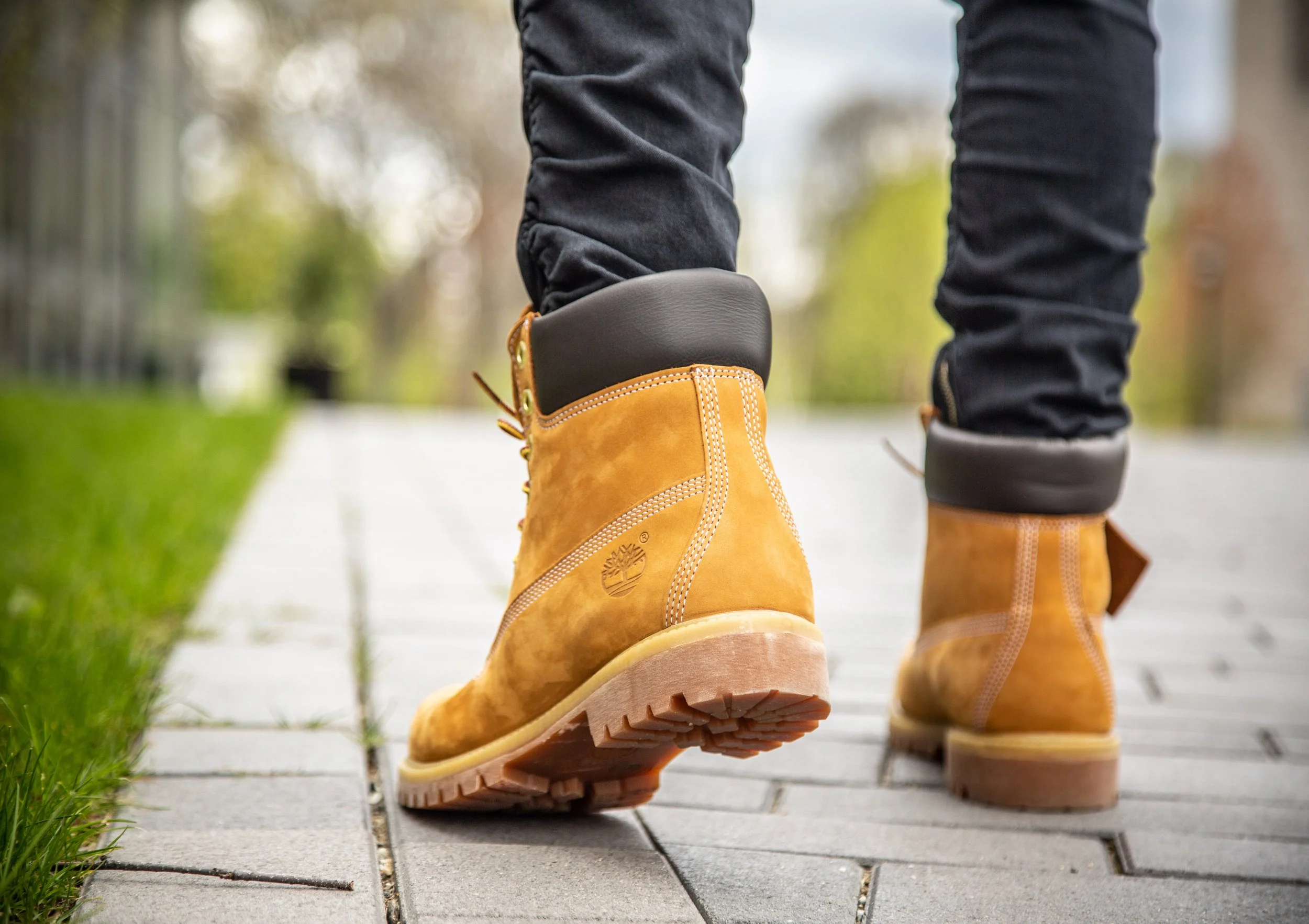 Close-up of a person walking on a pathway wearing tan Timberland boots and black pants, with green grass on the side and trees in the blurred background.