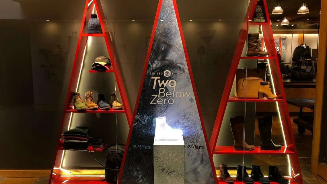 Display shelves shaped like red triangles holding Timberland boots, hats, and scarves in a retail store. A poster in the center features a white boot with the text "Two Below Zero."