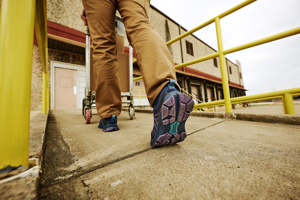 Person in tan pants and purple shoes walking on ramp with yellow railings and a warehouse building in the background.