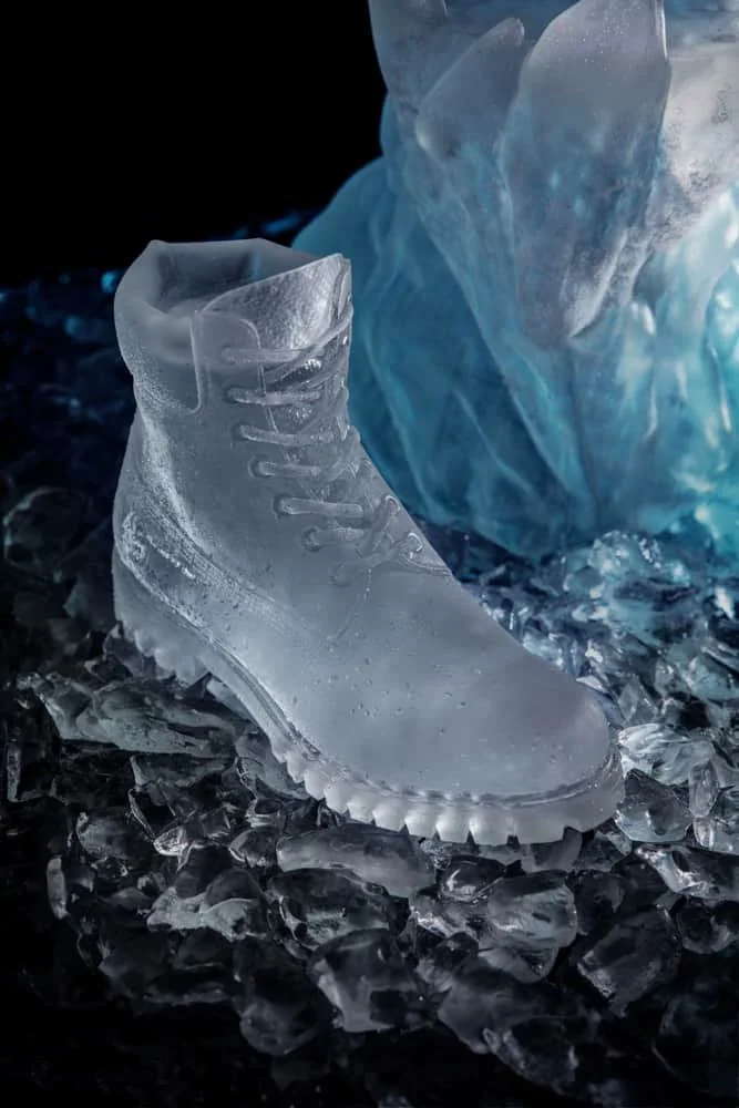 A transparent ice sculpture in the shape of a Timberland 6" boot, placed on ice shards with a blue ice sculpture in the background.