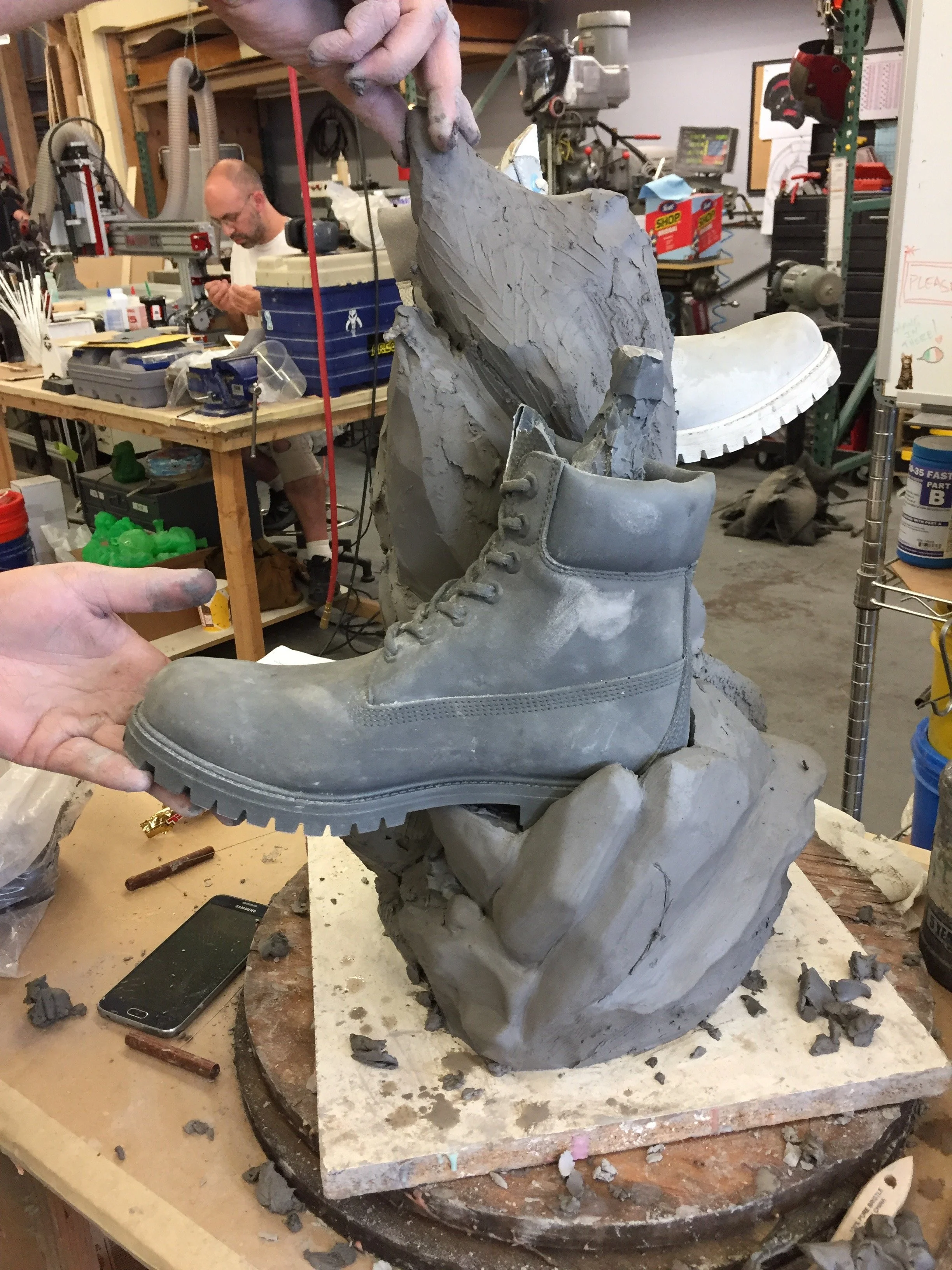 A person holds up a gray boot sculpture in a cluttered workshop with tools and supplies, with another person working in the background.