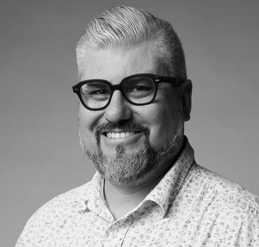 Black and white portrait of a smiling man with light-colored hair, beard, and glasses, wearing a patterned shirt against a plain background.