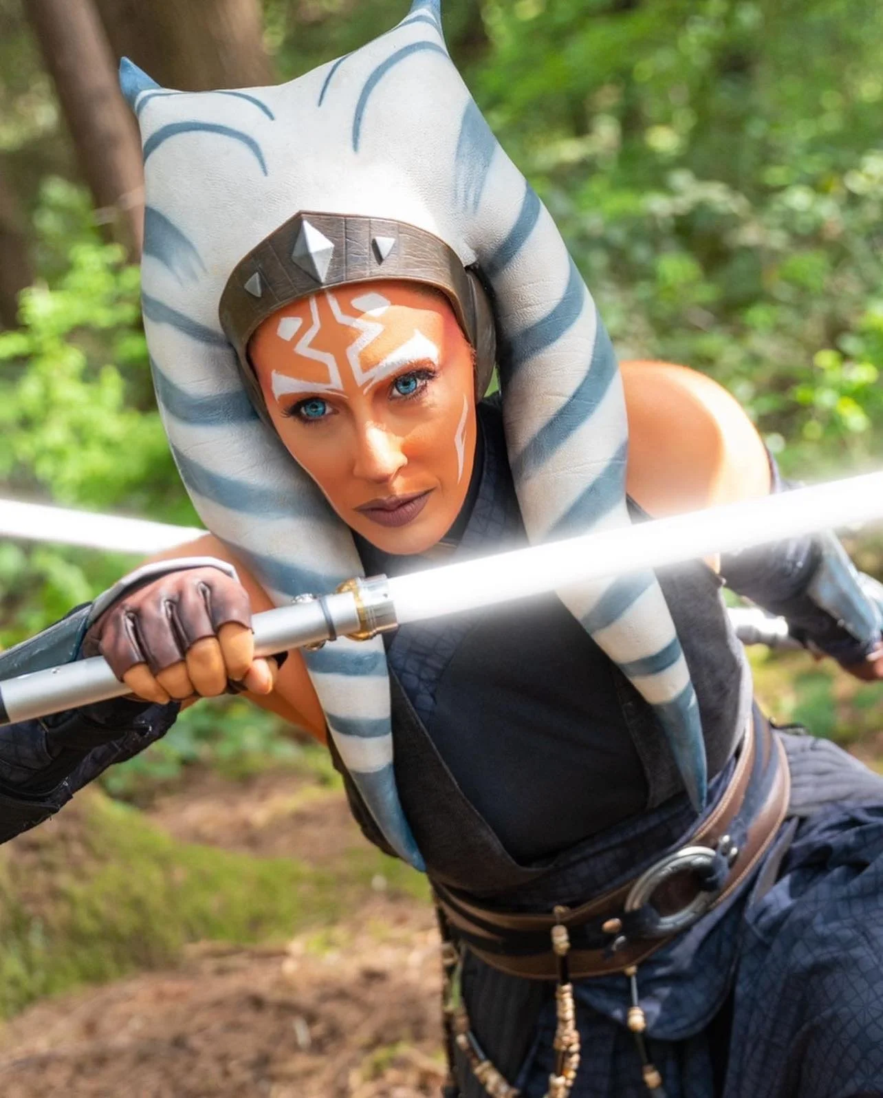 A woman cosplaying as Ahsoka Tano from Star Wars, wearing special effects make up, wielding a lightsaber, in a forest.