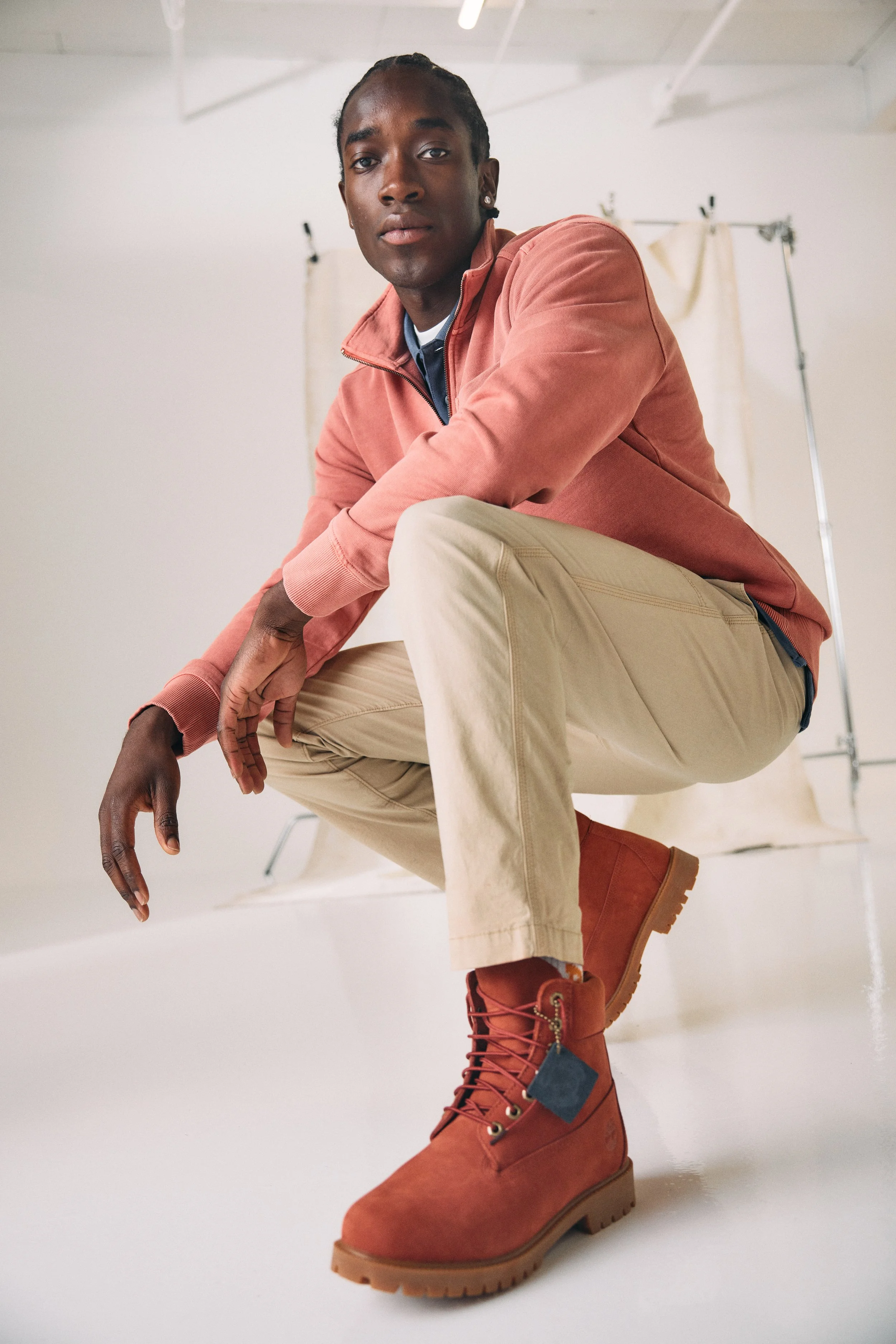 A person crouching and looking at the camera, wearing a pink shirt, tan pants, and red Timberland boots, with a neutral background and photography equipment in the background.
