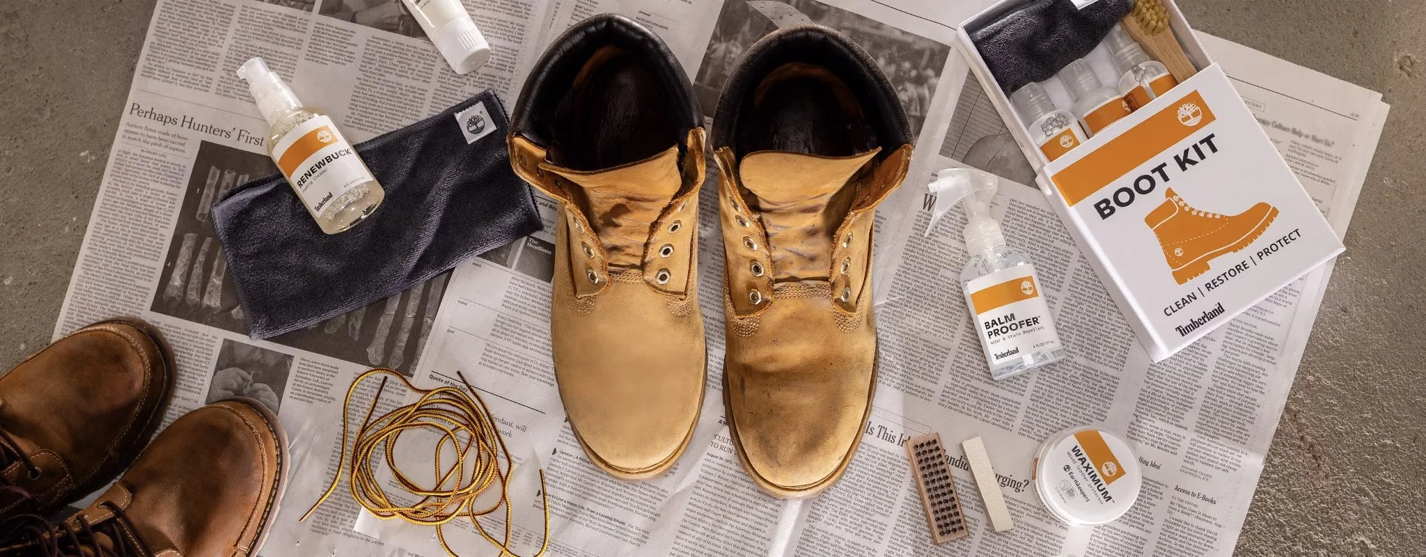 A pair of worn tan work boots, a bottle of Timberland boot cleaner, a spray bottle of balm proofer, a container of WAXIUM, a black cloth, a pair of brown shoes, yellow shoelaces, a nail file, and a boot restoration kit with small bottles and brushes,