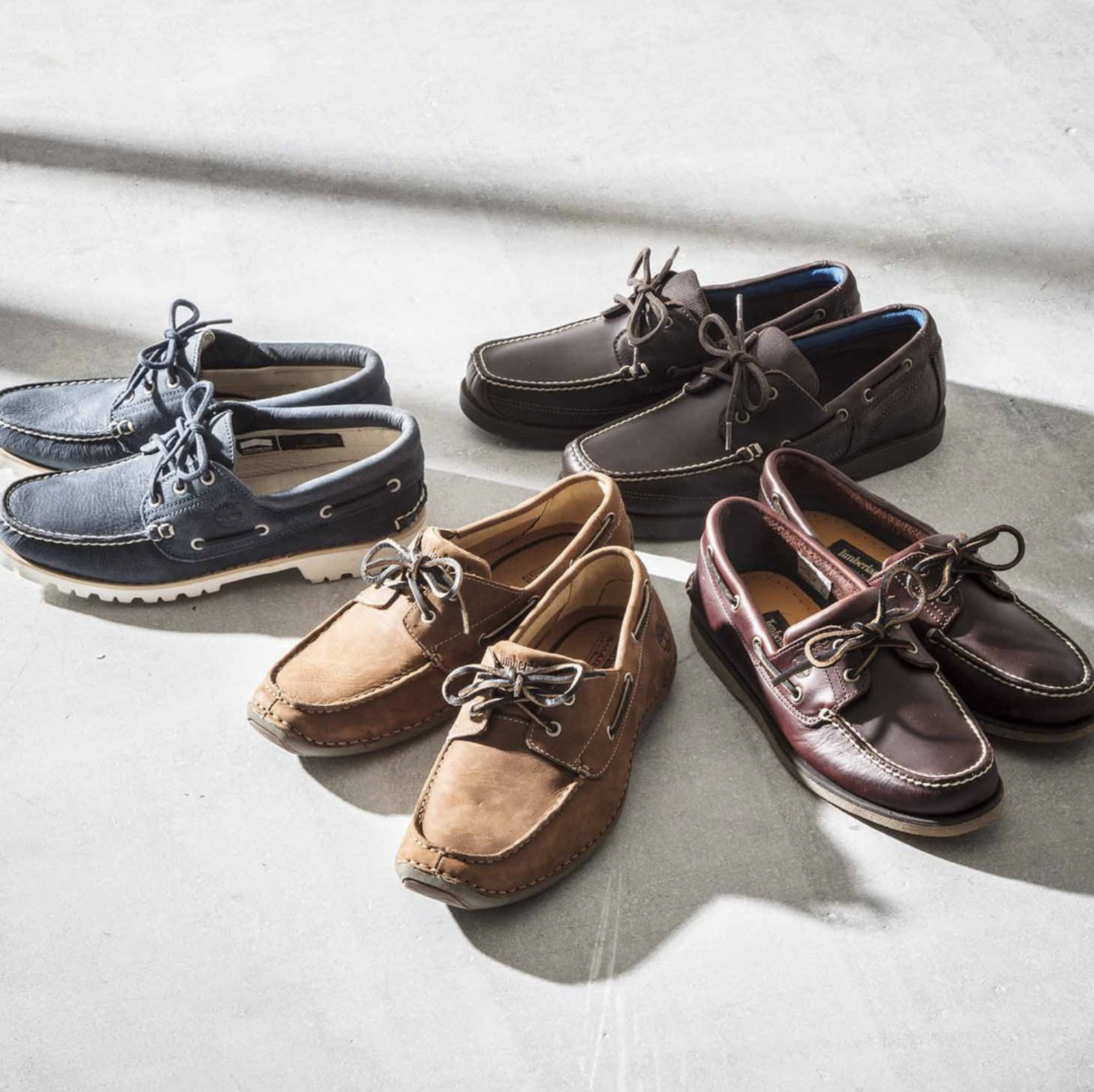 Six pairs of men's Timberland boat shoes arranged on a concrete floor, with sunlight casting shadows.