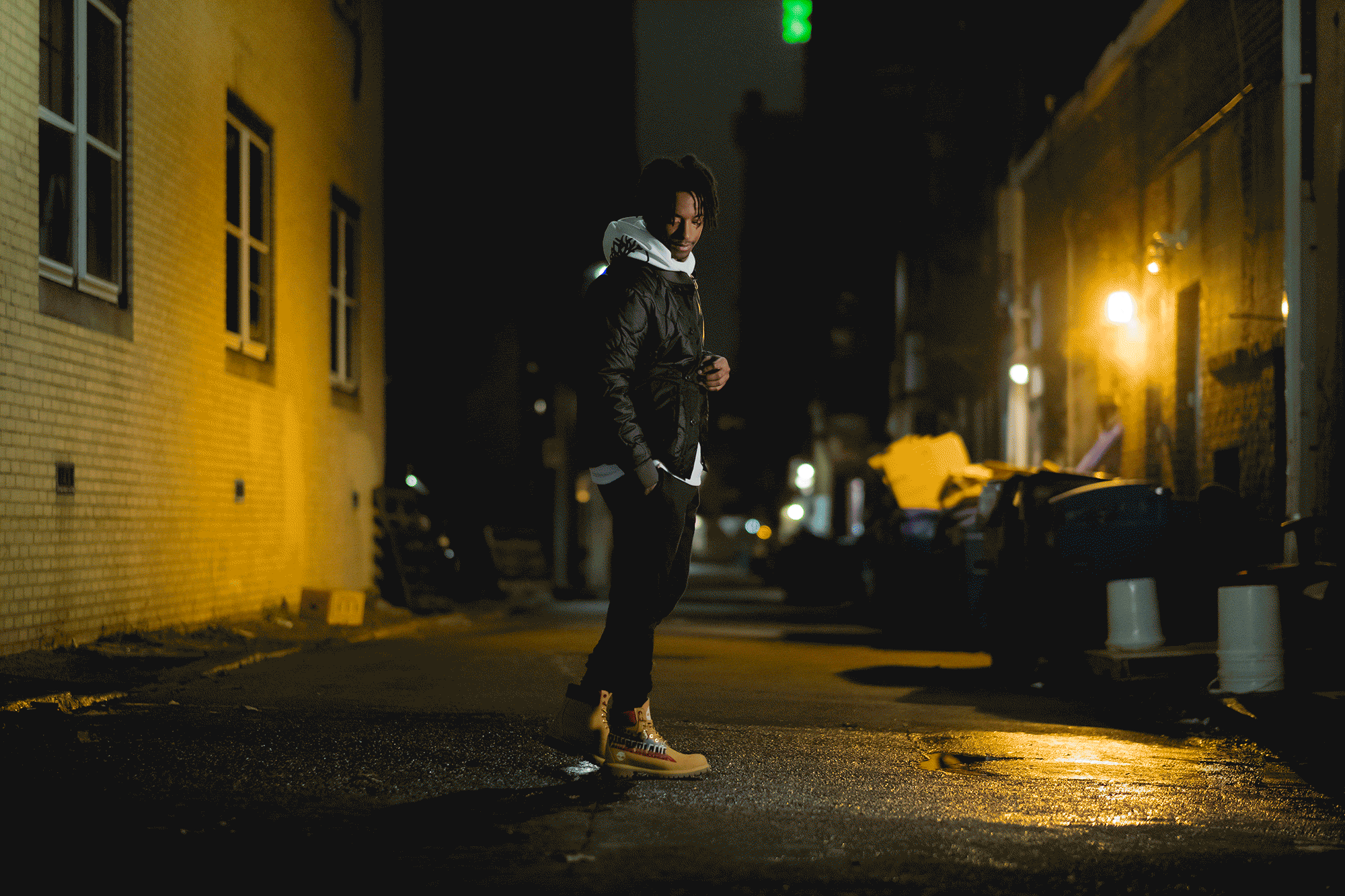 A person standing alone on a dimly lit street at night, wearing a black jacket, beige work boots, and a white hoodie. The street has yellow brick buildings and trash bins, illuminated by warm streetlights, with a wet pavement reflecting the lights.