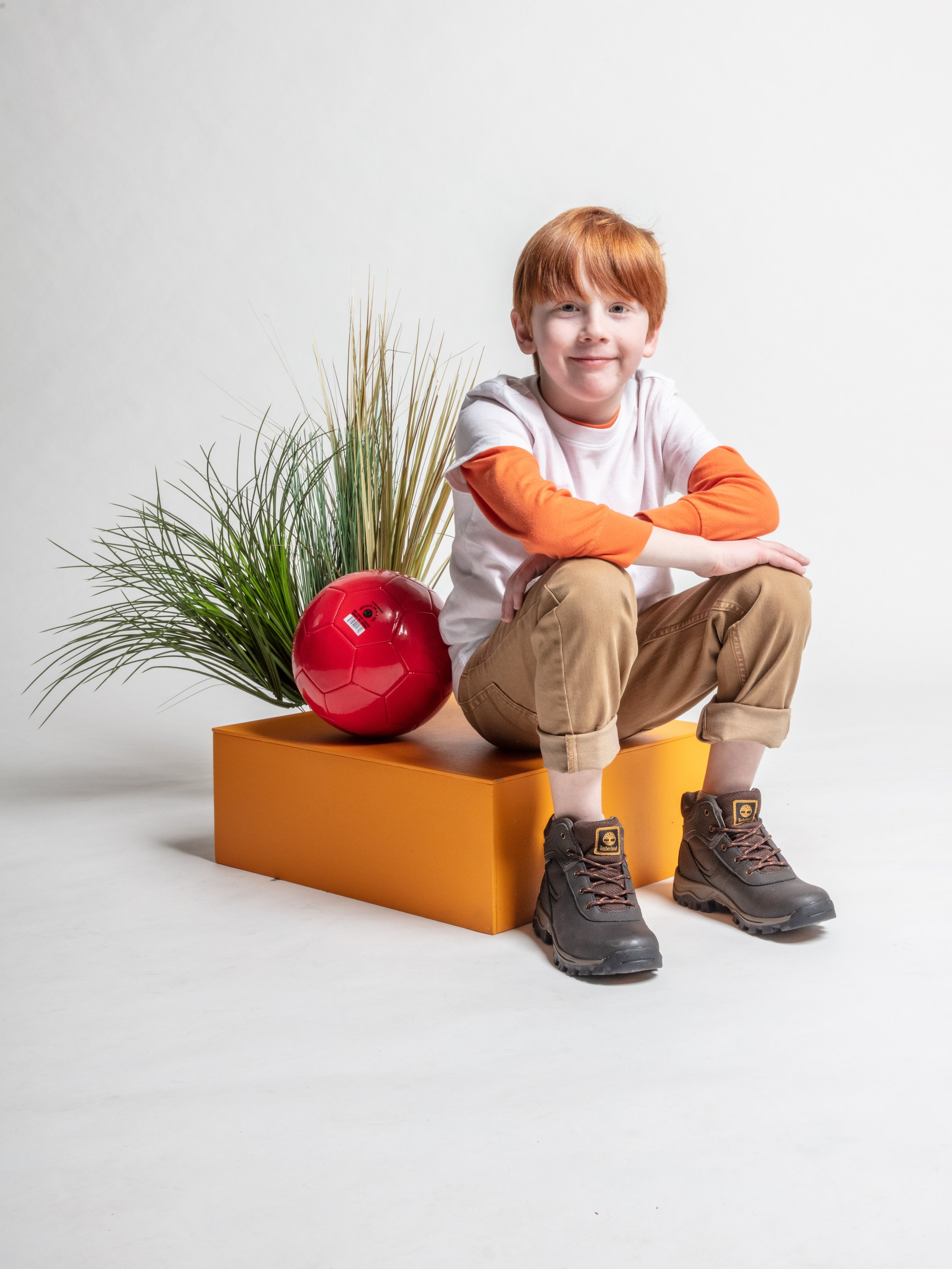 Young boy with red hair sitting on a yellow block, wearing boots, khaki pants, and a white shirt with orange sleeves, next to a red soccer ball and green plants, against a plain white background.
