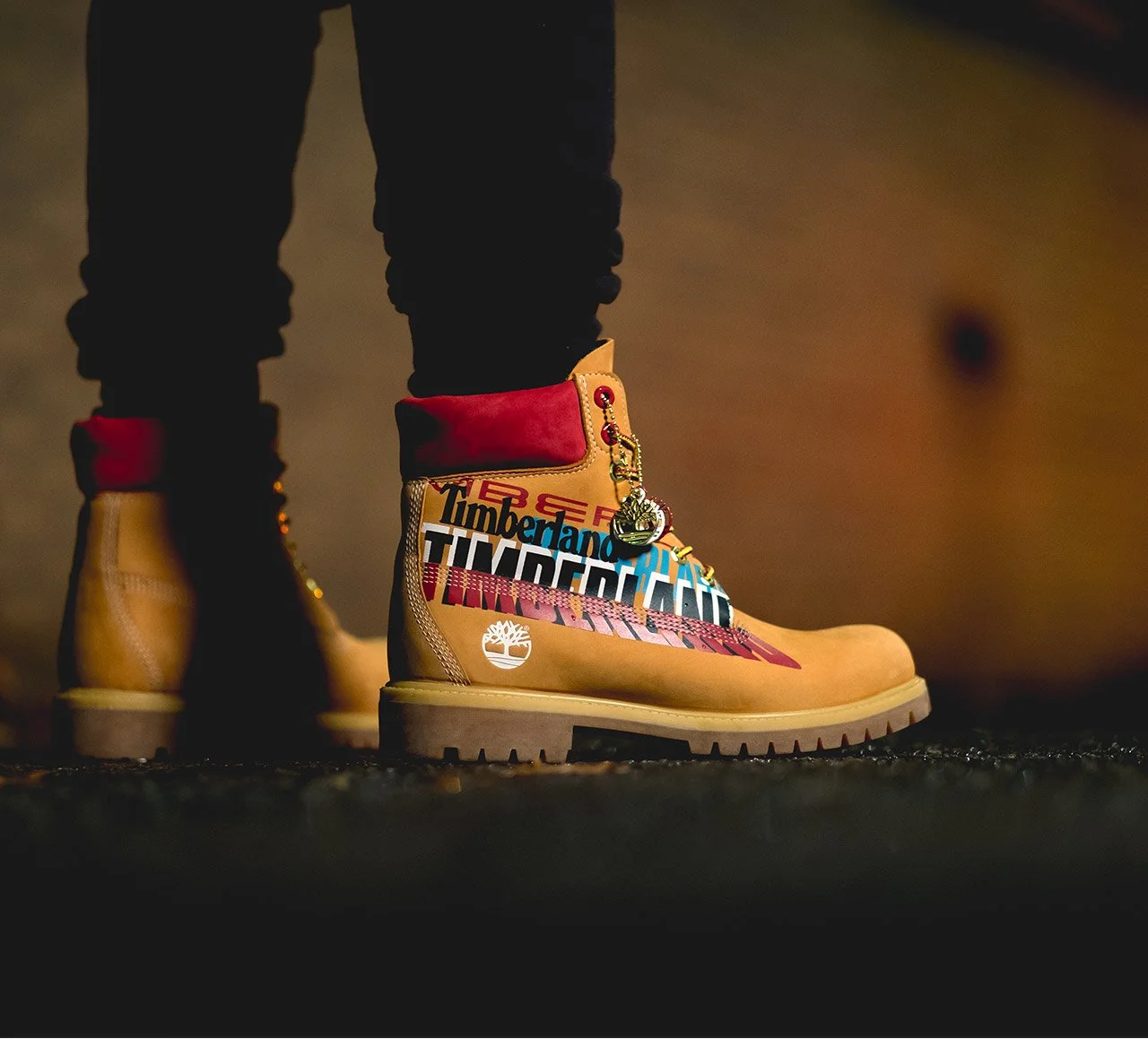 Close-up of tan Timberland boots with colorful logos and a red ankle collar, worn by a person standing on a dark surface, with the background showing part of a wooden surface.