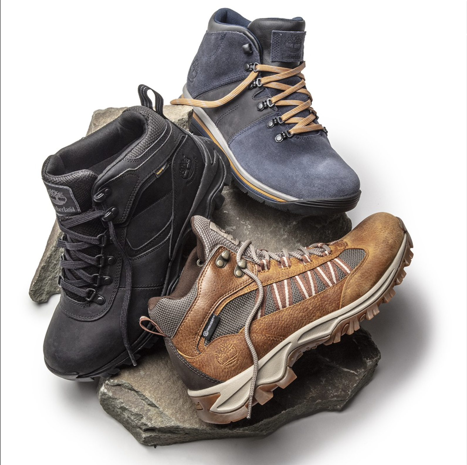 Three hiking boots of different styles and colors are displayed on a rock, with a white background.