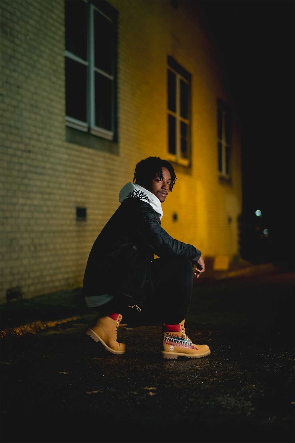 A young man with dreadlocks squatting on the pavement at night, wearing a black jacket with a white hood, black pants, and Timberland boots with a colorful design.