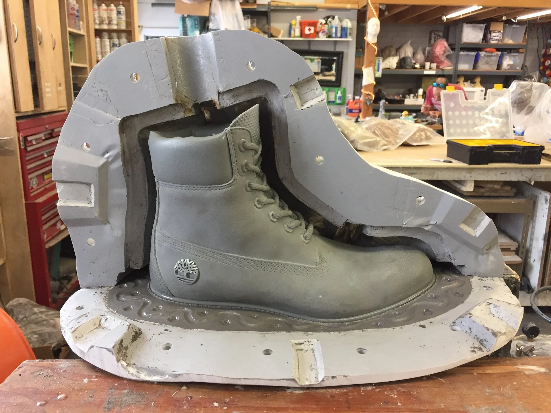 Timberland boot embedded in a mold for casting, with workshop tools and supplies in the background.