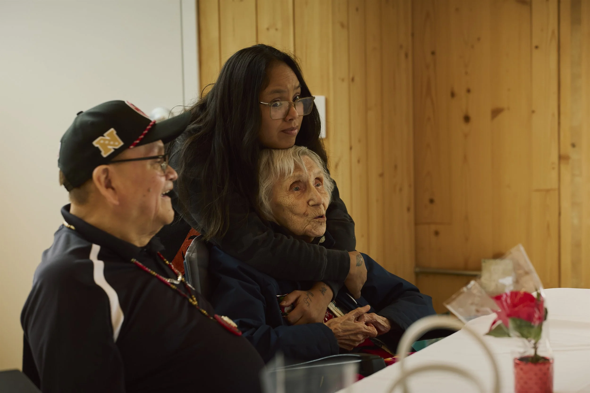 Three people sitting at a table, one elderly woman with gray hair, a woman with long dark hair and glasses, and a man wearing a black cap, celebrating a special occasion with flowers and possibly a cake.