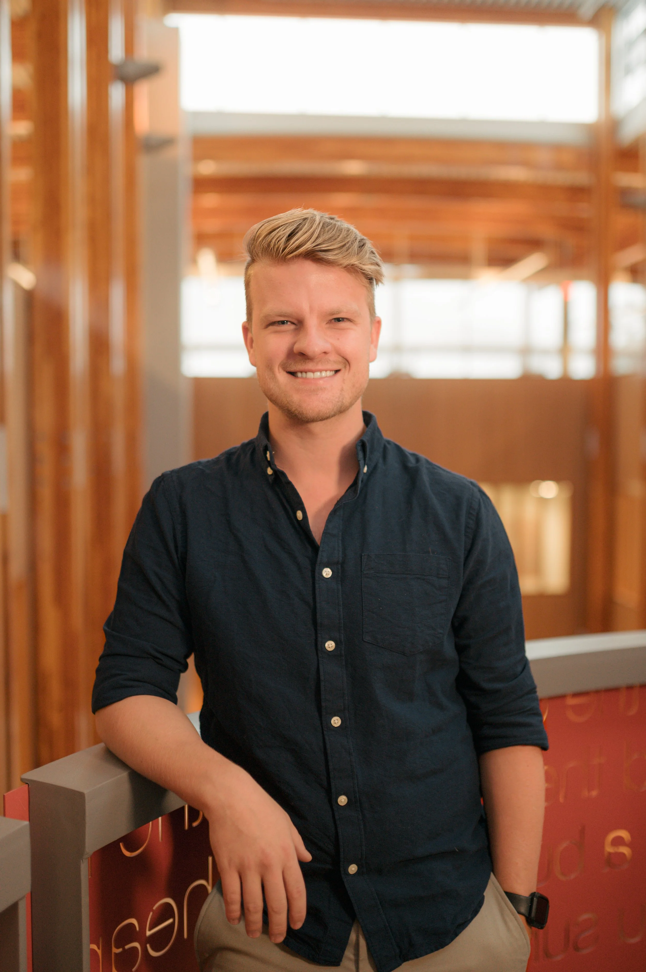 A young man with blonde hair, wearing a dark button-up shirt and khaki pants, smiling and leaning on a railing inside a building with wooden interior design.