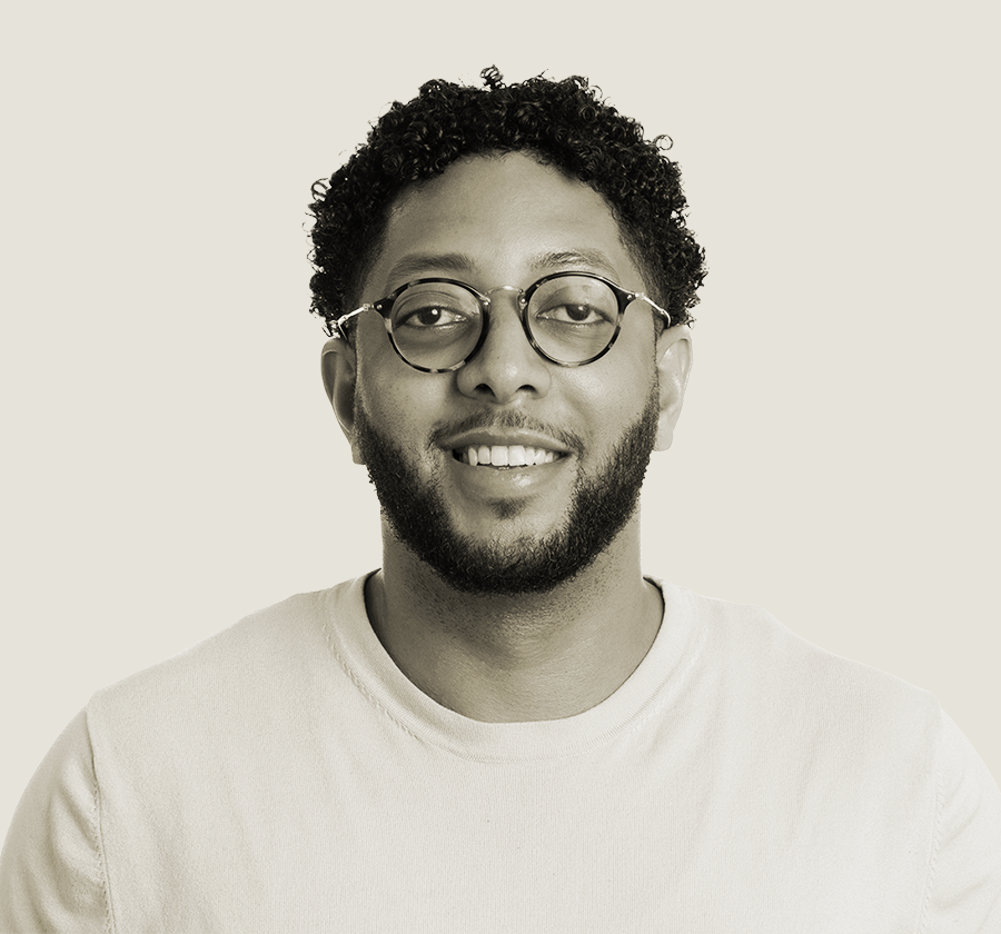 A portrait of a young man with curly hair, glasses, and a beard, smiling against a plain background.