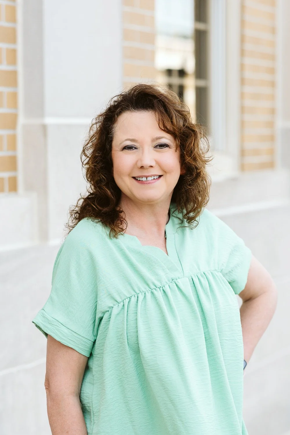 A smiling woman with light brown, curly hair, wearing makeup, earrings, and a black top, against a gray background.
