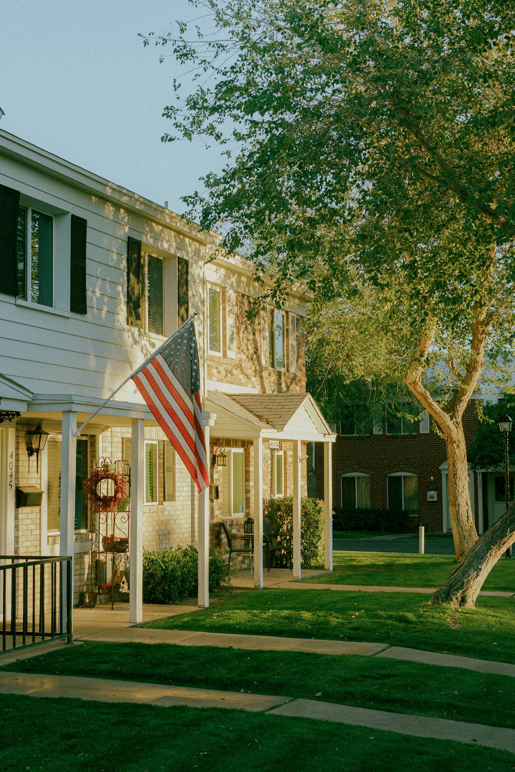 A row of suburban houses with front porches, one with an American flag, and a large tree in the front yard. The scene is illuminated by warm sunlight.