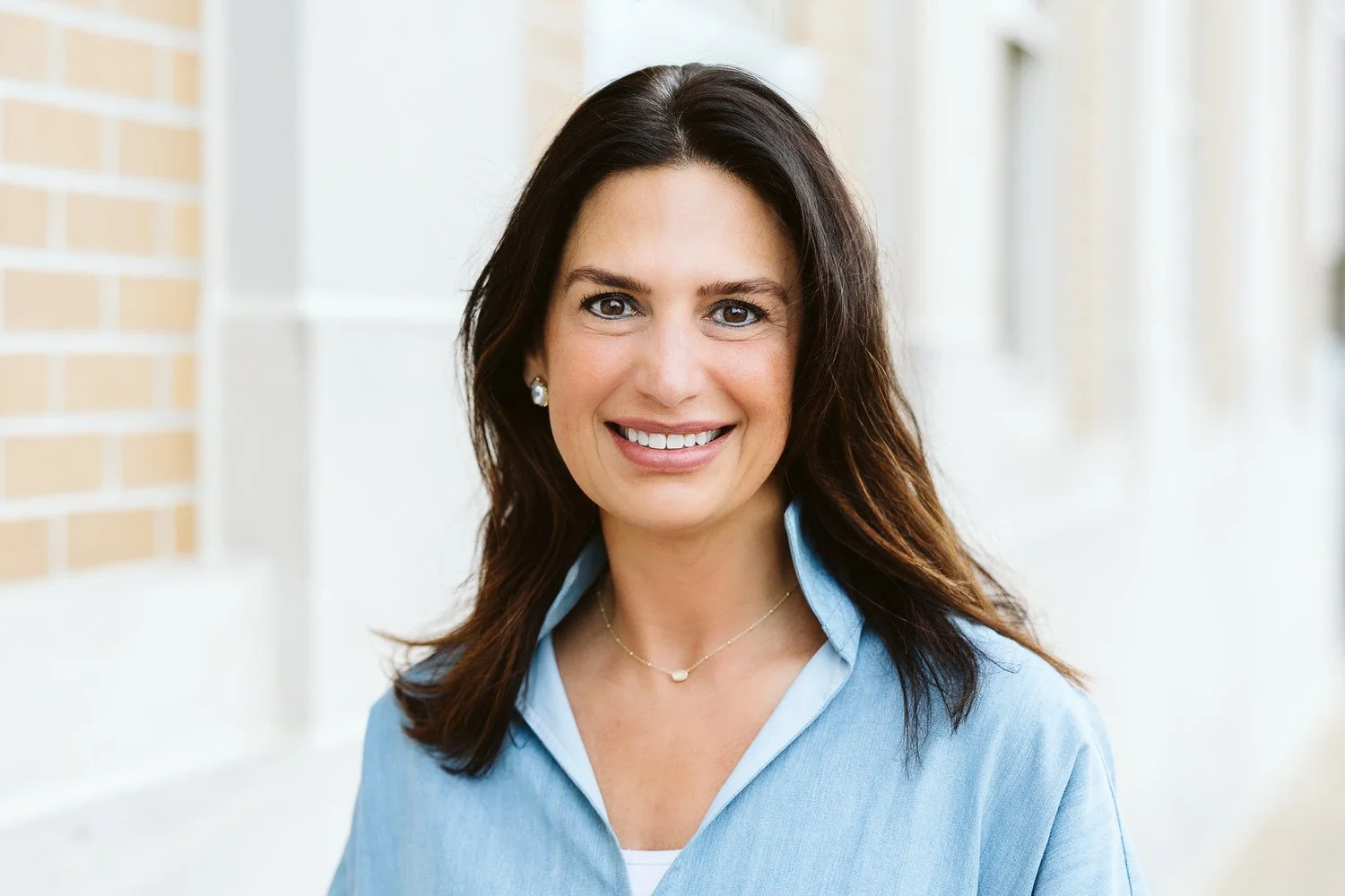 A smiling woman with long brown hair, wearing a light blue button-up shirt, standing outdoors with a brick wall and windows in the background.