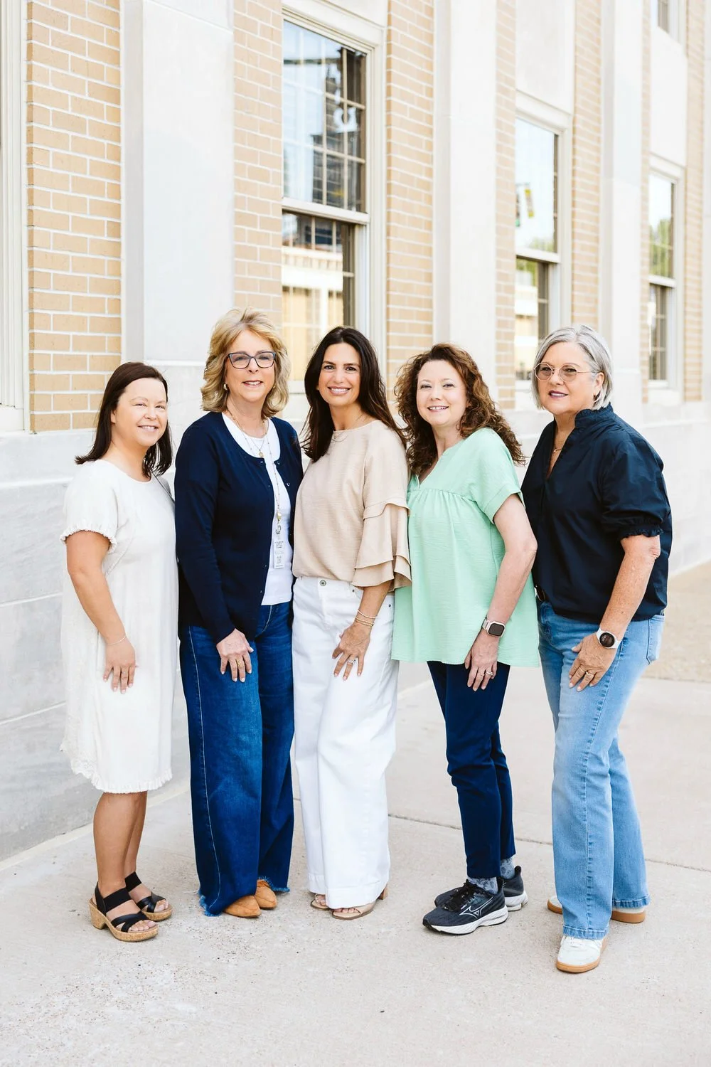 Five women standing outside in front of a brick building, smiling at the camera.