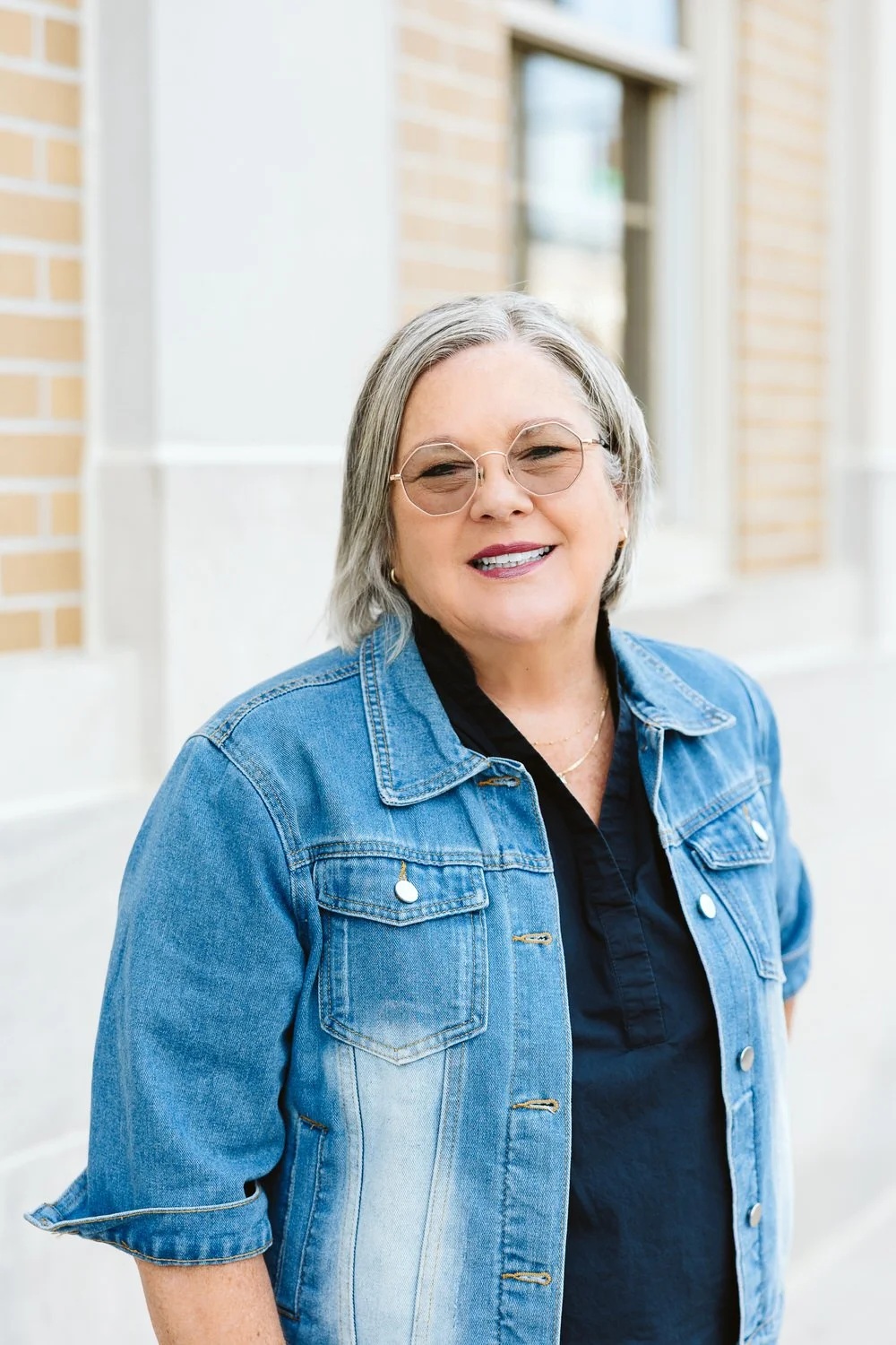 Close-up of a middle-aged woman with gray hair, wearing glasses, a black top, and smiling against a gray background.