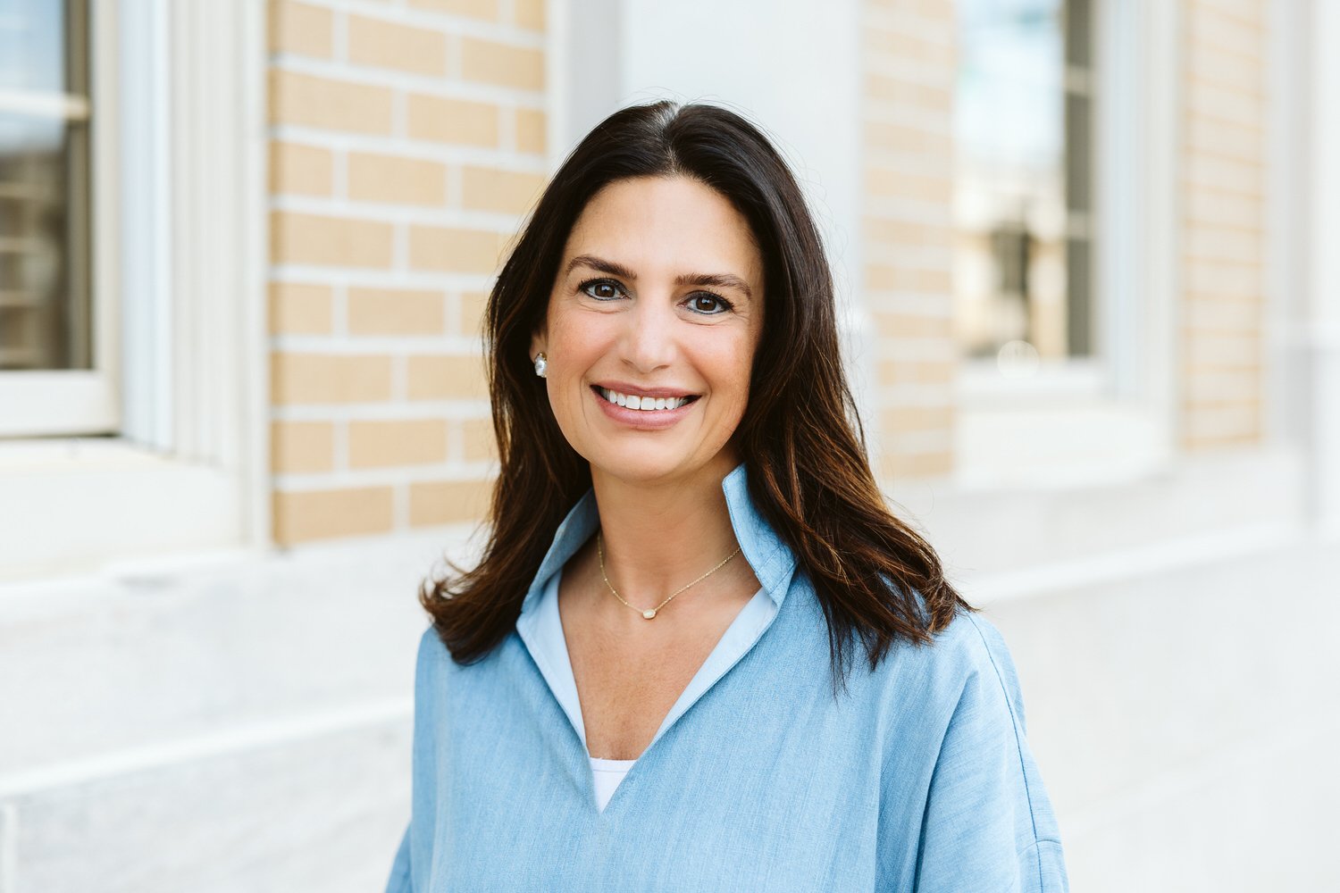 A woman with shoulder-length dark brown hair smiling outdoors near a building with brick walls and windows, wearing a light blue collared shirt and jewelry.