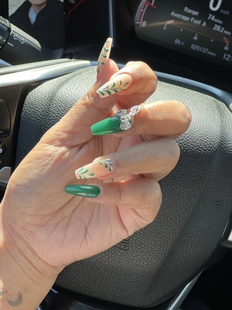 Close-up of a hand with long, decorated nails in green and beige with leaf designs, wearing a silver diamond ring, held in front of the steering wheel of a car with a digital dashboard display.