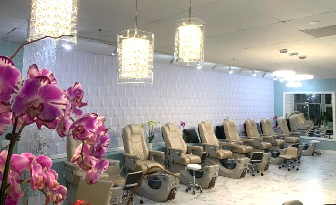 Nail salon with multiple beige pedicure chairs, pink orchids in the foreground, and modern chandeliers hanging from the ceiling.