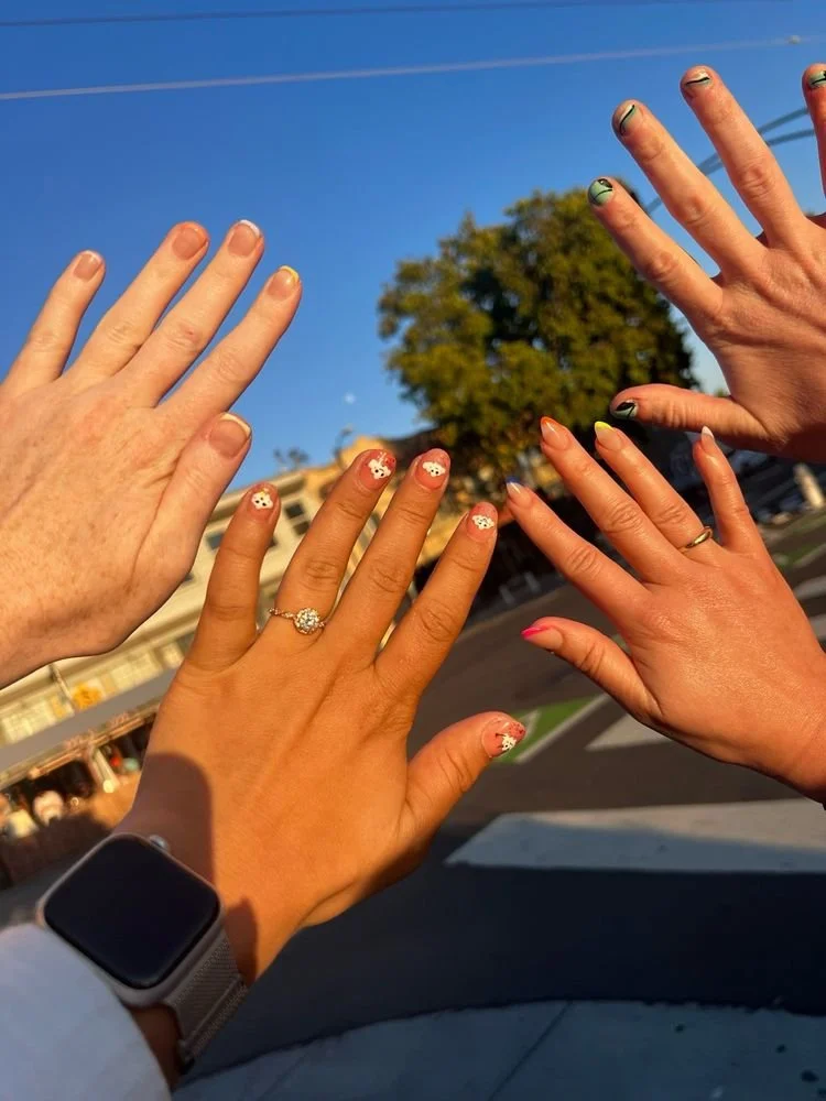 Four hands with painted nails and rings extended upward outdoors, against a clear blue sky with a tree and buildings in the background.